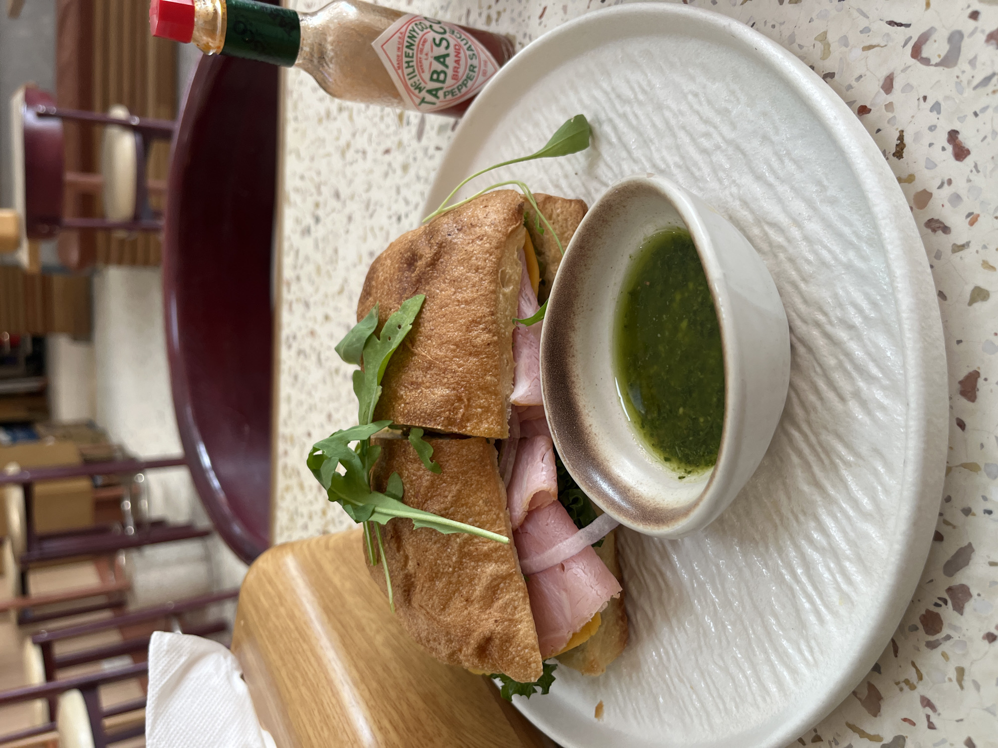 A high-angle shot shows a cut ham sandwich on a textured white plate next to a small bowl of green sauce and a bottle of Tabasco sauce. The sandwich, cut in half, is filled with ham, cheese, greens, and sliced onion. The white plate has a textured, ridged pattern. The small bowl contains a vibrant green sauce. A bottle of Tabasco hot sauce is placed above the sandwich. The plate rests on a surface with a speckled, terrazzo-like design. The background is slightly blurred but suggests a cafe or restaurant setting, with furniture partially visible. Based on the prompt context, this is assumed to be located in Bac Nha Trang Ward, Vietnam.
