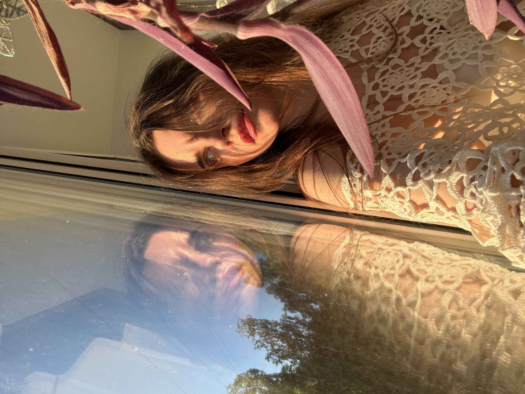 A close-up, rotated portrait captures a young woman illuminated by golden light, likely taken indoors near a window in Chaikovsky, Russia. The woman's head and upper torso are visible, positioned horizontally across the upper half of the frame when oriented correctly. She has long, flowing brown hair, and her left eye, light in color, is open, looking towards the viewer with a neutral expression. Her lips are slightly parted, showcasing a glossy, pink hue.
A plant with striking deep purple or maroon leaves frames the foreground, partially obscuring parts of her hair and face, adding a natural element to the composition. She is wearing a light-colored, possibly cream or white, crocheted garment with an intricate open-work pattern visible on her shoulder and chest.
A significant feature of the image is the reflection on what appears to be a window or glass surface. Her face and part of her upper body are reflected, creating a layered visual effect. The reflection also reveals the outdoor scene: a clear blue sky, green trees, and faint indications of power lines or wires in the distance. The warm, golden quality of the light strongly suggests the photograph was taken during the "golden hour," either early morning or late afternoon, on a sunny day. No visible text is present in the image.