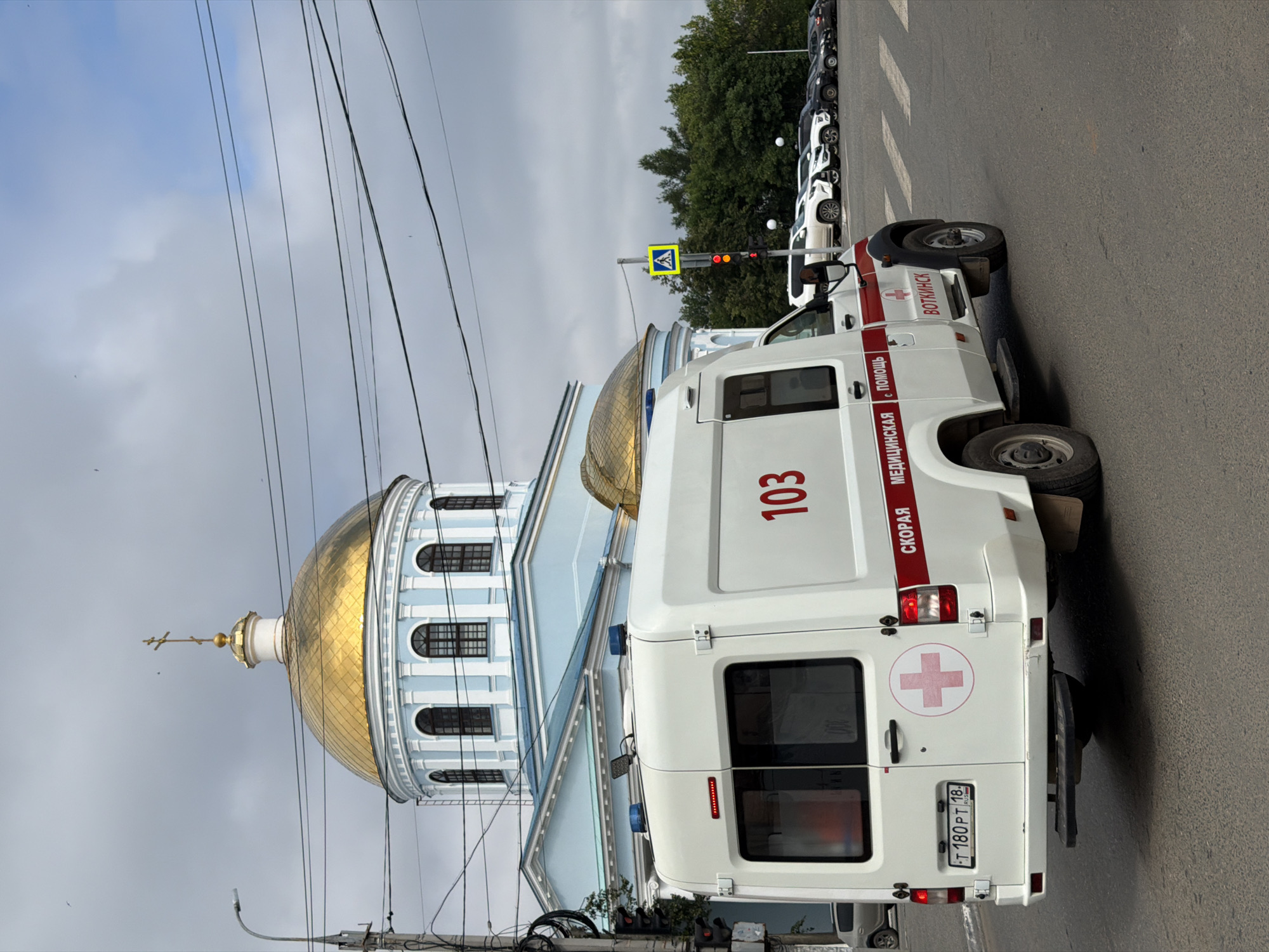 An ambulance, marked with "103" and "СКОРАЯ МЕДИЦИНСКАЯ ПОМОЩЬ" (Emergency Medical Aid), is seen from a rear, right-side perspective, driving on a paved road in the city of Votkinsk, Russia. The ambulance, a white van, has red stripes and a red cross emblem. Behind the ambulance, a light blue building with a prominent golden dome, characteristic of a church or cathedral, is visible. Several cars are parked in a lot to the right of the ambulance. Overhead, power lines crisscross the sky, which is overcast with patches of blue. A traffic light and a pedestrian crossing sign are also present. The overall scene depicts a street view in Votkinsk with an ambulance in transit, set against the backdrop of religious architecture.