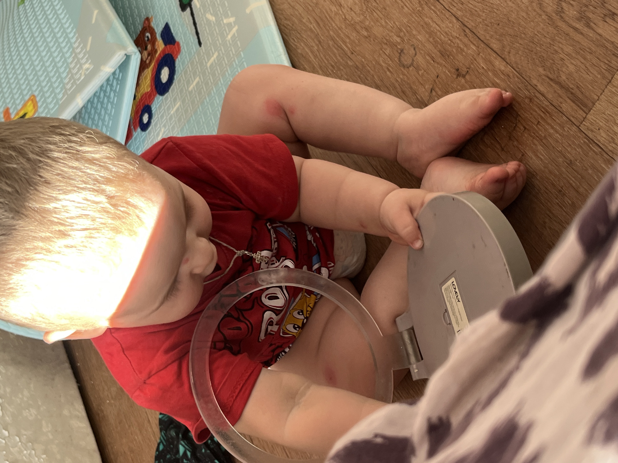 A toddler in a red t-shirt with cartoon characters is playing on a wooden floor. They are sitting next to a blue and white play mat with images of vehicles. The toddler is holding a round, gray object with a transparent ring. Their legs are visible, and there appears to be a red mark on their knee. A beam of sunlight is shining on the toddler's head. A tag on the gray object reads "LOVELY". The setting appears to be indoors.