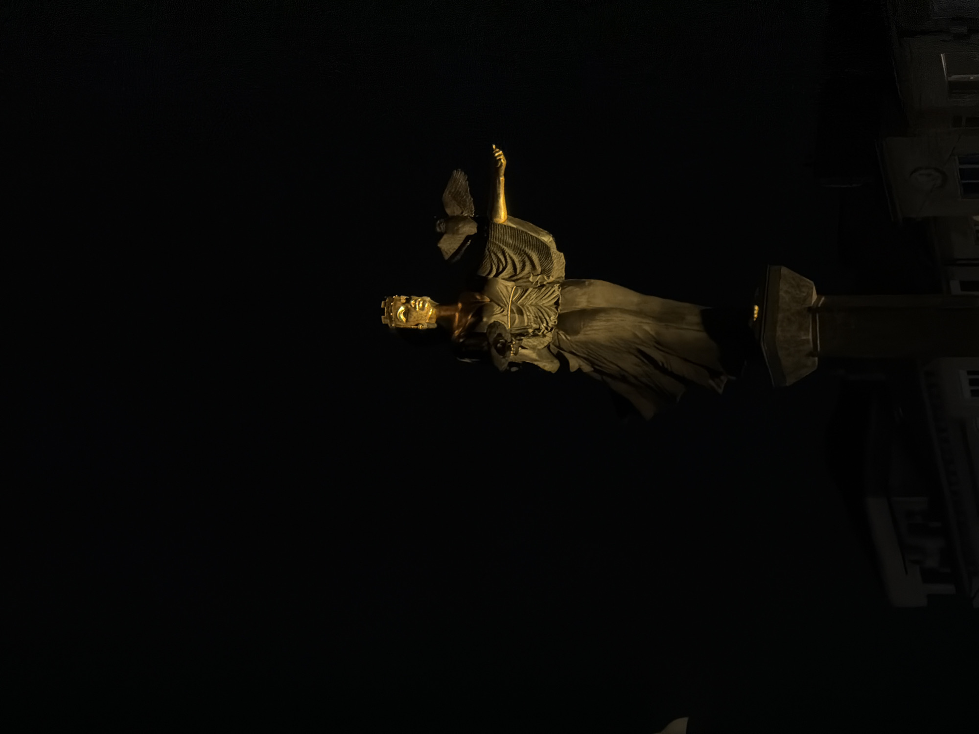 The image shows the Sofia Justice Palace at night, in Sofia, Bulgaria. The statue is illuminated by a golden light and stands in sharp contrast to the surrounding darkness. The statue appears to be a woman with one hand raised. The Justice Palace building is visible in the background, dimly lit.