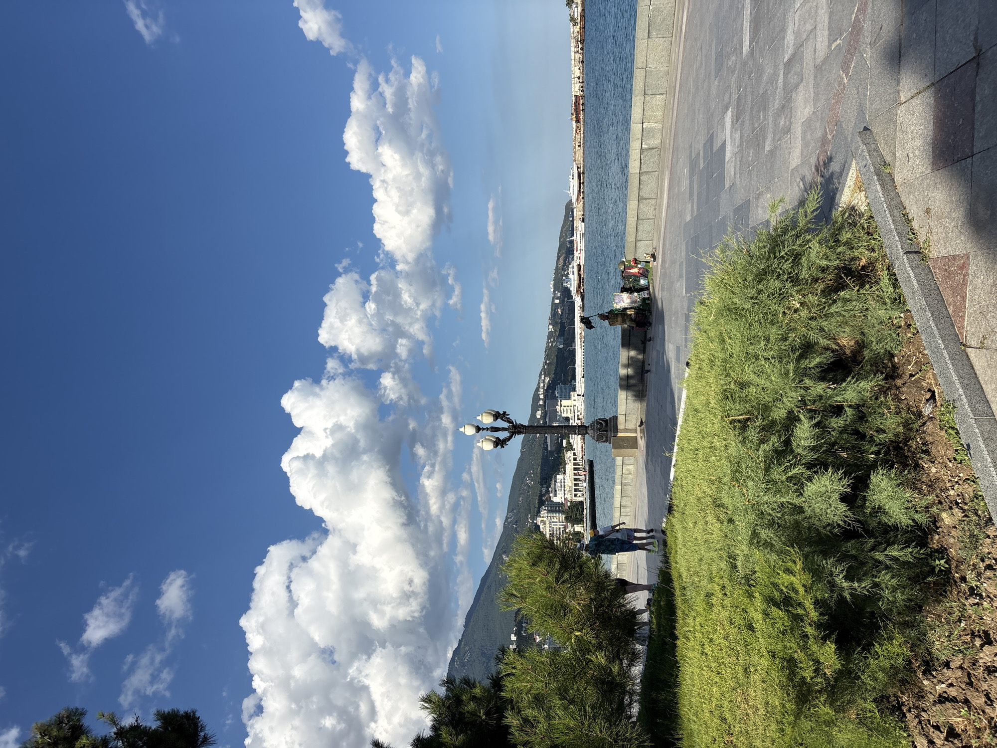 The image shows a seaside promenade in Yalta, Ukraine, on a clear day with scattered white clouds against a bright blue sky. In the foreground, a lush green bush occupies the lower left corner, bordering a paved walkway. The walkway is made of grey stone tiles with a reddish accent pattern. To the right of the walkway, the blue sea stretches out, with several white boats docked in the distance.
Along the promenade, a black lamppost with three ornate white lights stands prominently. A stone wall borders the promenade and the sea. In the mid-ground, a couple walks along the promenade, facing away from the camera. Further down the promenade, near the sea wall, a person sits on a small scooter or cart, surrounded by various items, possibly for sale or display, including a black flag. Pigeons are visible on the ground near the seated individual.
In the background, a range of buildings, some white and modern, others with a more classical appearance, line the coastline. Behind the buildings, a large, green mountain rises, its slopes dotted with smaller structures. The overall scene depicts a vibrant coastal city setting, with a mix of natural beauty and urban development.