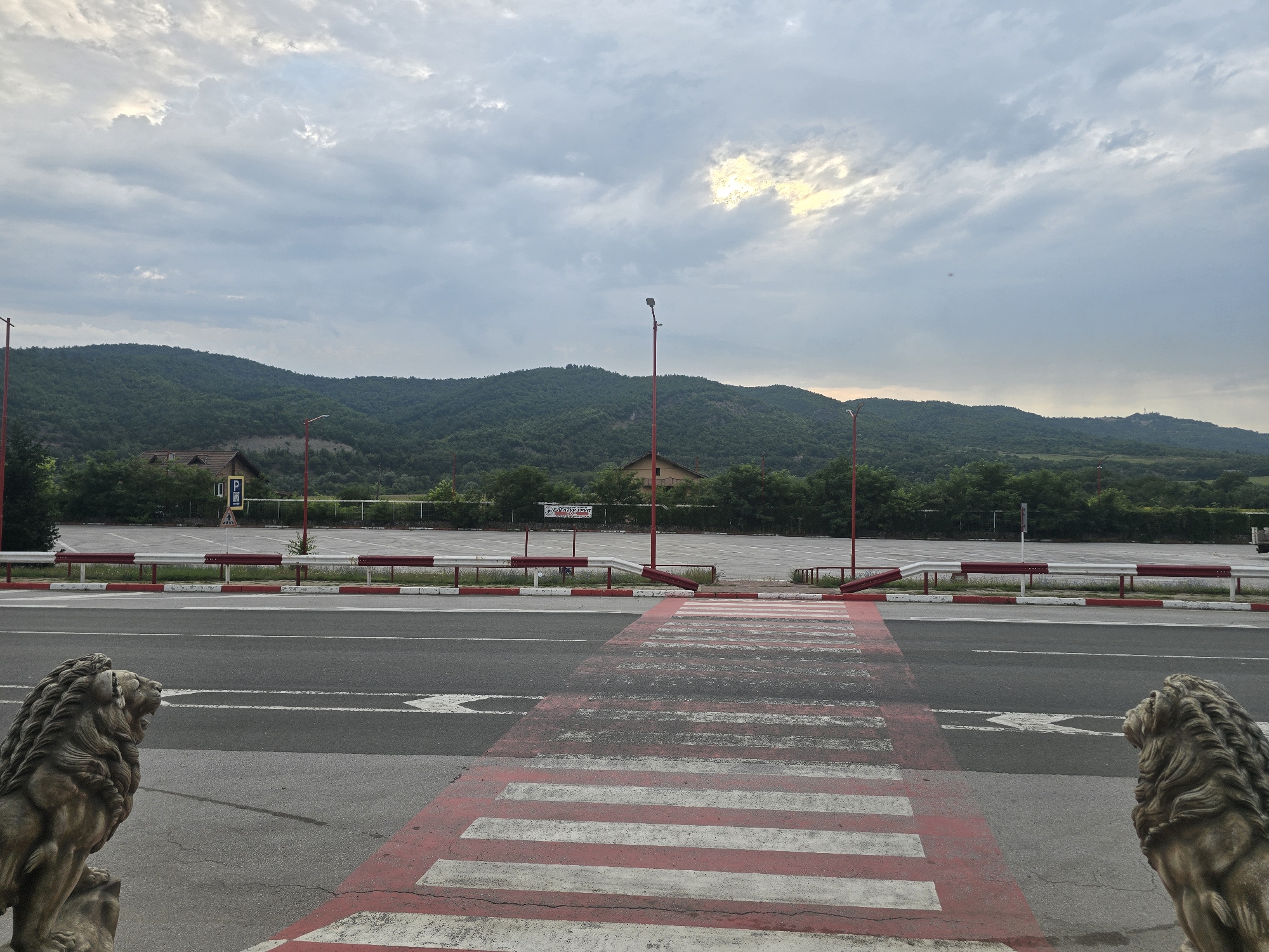 The image depicts an outdoor scene with a focus on a road and a parking lot, set against a backdrop of rolling, tree-covered hills. In the foreground, a pedestrian crossing with red and white stripes is visible, leading across a grey asphalt road. On either side of the crossing, stone lion statues are placed. The road has lane markings. Beyond the road, a white barrier with red accents separates it from a large, empty asphalt parking lot. Several red-topped lampposts are positioned throughout the scene, with one prominently in the middle ground. In the distance, a few buildings and dense green foliage are visible at the base of the hills. The sky is overcast with patches of sunlight breaking through the clouds, suggesting a late afternoon or early morning setting. The image appears to be taken in Zlatna Panega, Bulgaria, given the context provided. There are no people or animals visible in the frame.