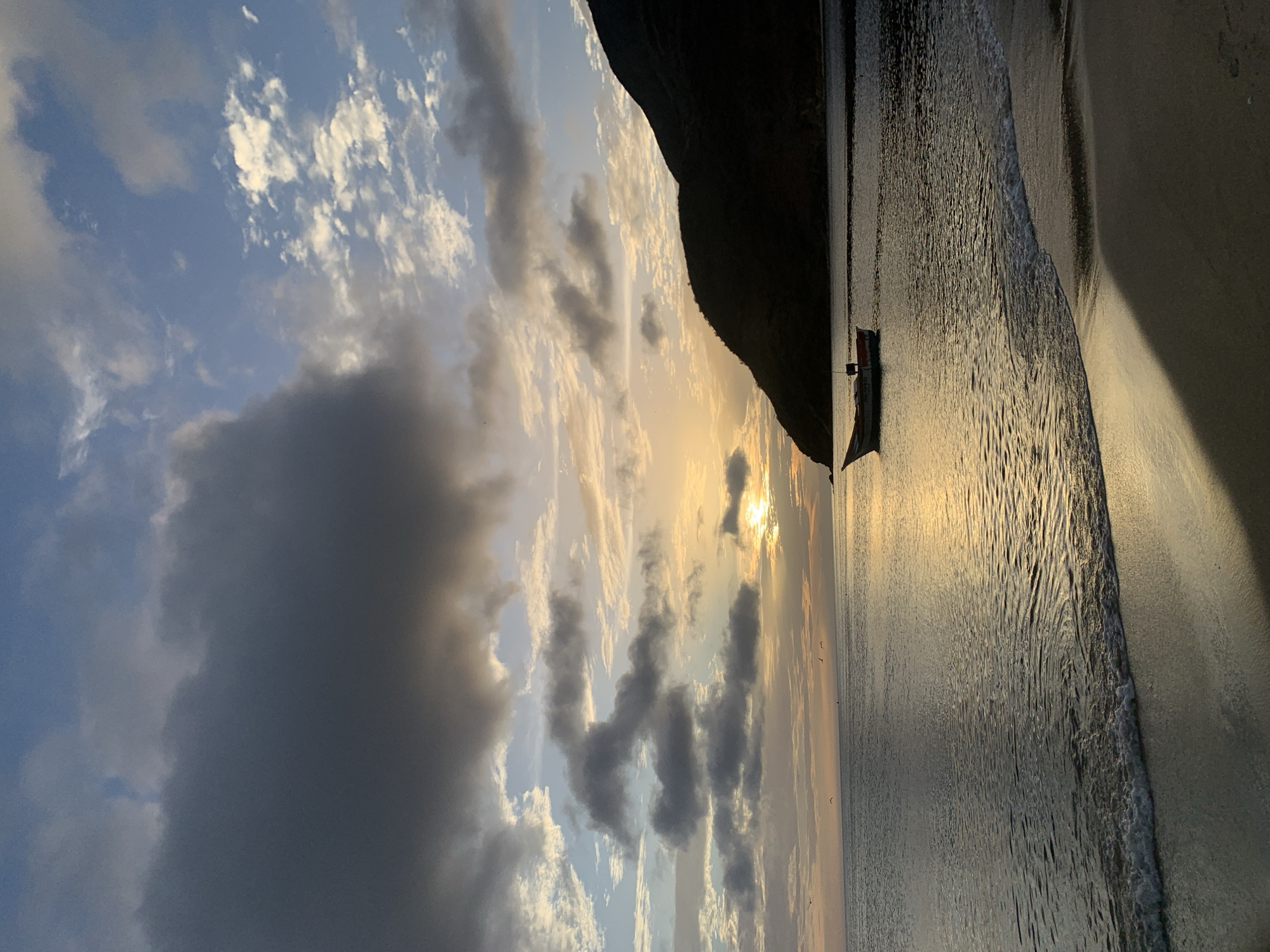 A small fishing boat is anchored in the calm waters of a beach in Juan Griego, Venezuela. The sun is setting behind a dark, silhouetted mountain, casting a golden glow on the water and sky. Clouds dot the sky, some dark and heavy, others wispy and light. The sand beach in the foreground is wet from the receding tide, with small waves gently lapping at the shore. The scene is peaceful and serene, with a sense of isolation and natural beauty.