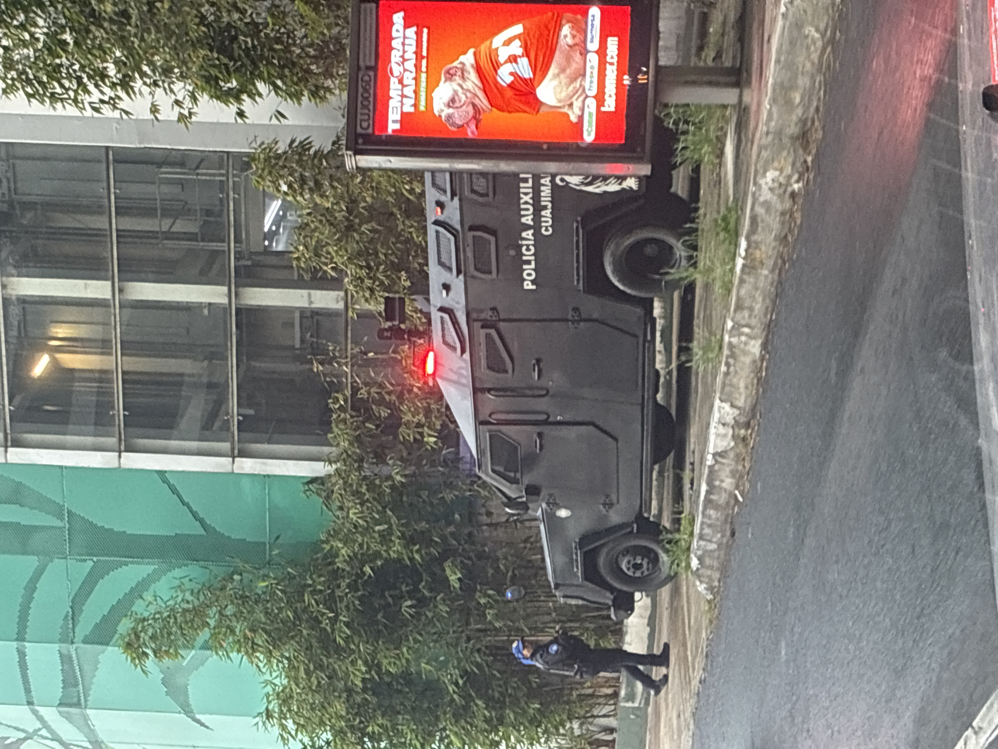 The image shows an armored police vehicle, identified by the inscription "POLICIA AUXILIAR CUAUJIMALPA" on its side. A police officer stands near the vehicle, which is parked on the side of the road. Above the vehicle is an advertising billboard. The environment appears urban, with trees and a building visible. The location is Cuajimalpa, a borough in Mexico City, Mexico.