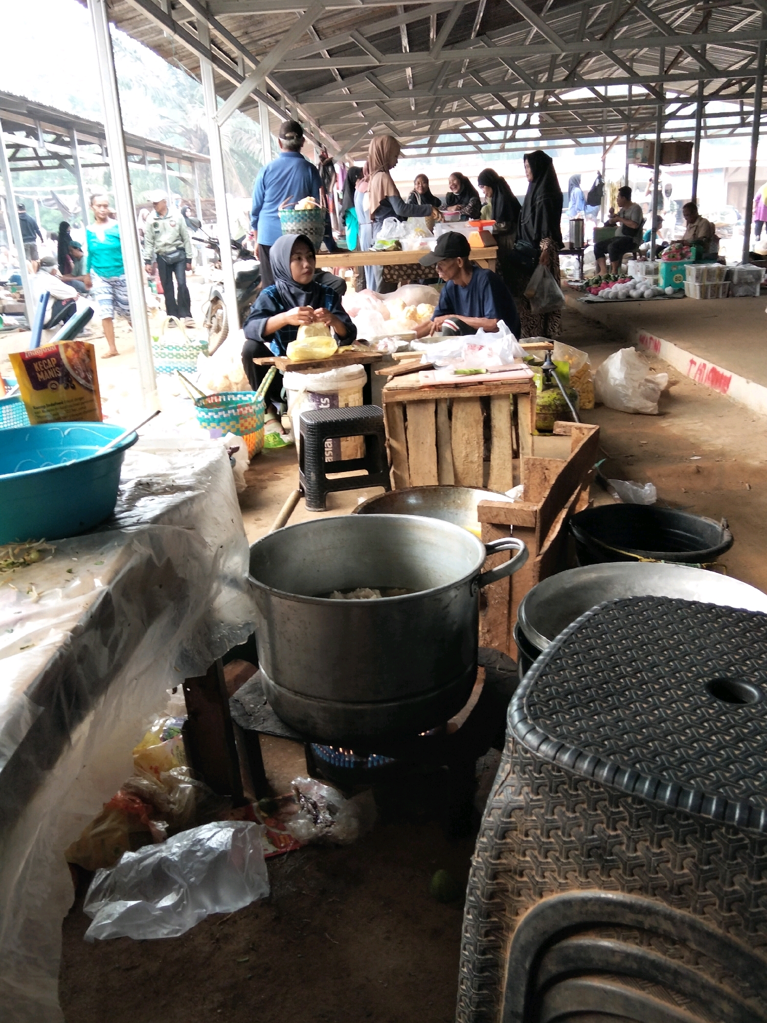 This is a medium-shot, eye-level, outdoor photograph of a bustling marketplace in Tirto Sari, Indonesia. The scene is filled with people engaged in various activities. In the foreground, a large, silver pot with a lid sits on a burner with a visible flame. To the right of the pot is a black, woven plastic stool. To the left of the pot is a blue plastic basin filled with vegetables. Behind the basin is a yellow and red packet labeled "Indofood Kecap Manis". Further back, a person wearing a dark headscarf and dark clothing is seated at a wooden table, appearing to be preparing food or packaging items. A variety of other individuals are visible in the background, including some wearing headscarves and others in casual attire. They are seen standing, walking, and interacting with each other. The marketplace appears to be under a covered structure with a corrugated metal roof supported by metal beams and wooden poles. Various wooden stalls and tables are set up, displaying goods and serving as work surfaces. The ground is dirt and concrete. The overall impression is one of a busy, functional marketplace where daily commerce is taking place.