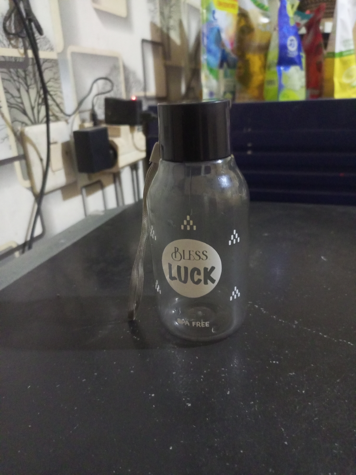A clear plastic water bottle with a black lid and a white label featuring "BLESS LUCK" and "BPA FREE" is positioned on a dark, speckled surface. The bottle has a thin string attached to its neck. In the background, out of focus, are several bags of what appear to be cleaning supplies or food items in various colors, including blue, yellow, and red. To the left of the bottle, also out of focus, is a wall with framed pictures and electrical cords connected to a black power adapter with a small red light. The lighting is dim, casting some shadows. The scene appears to be indoors, possibly in a residential setting. There is no visible indication of Cidahu, Indonesia in the image itself.