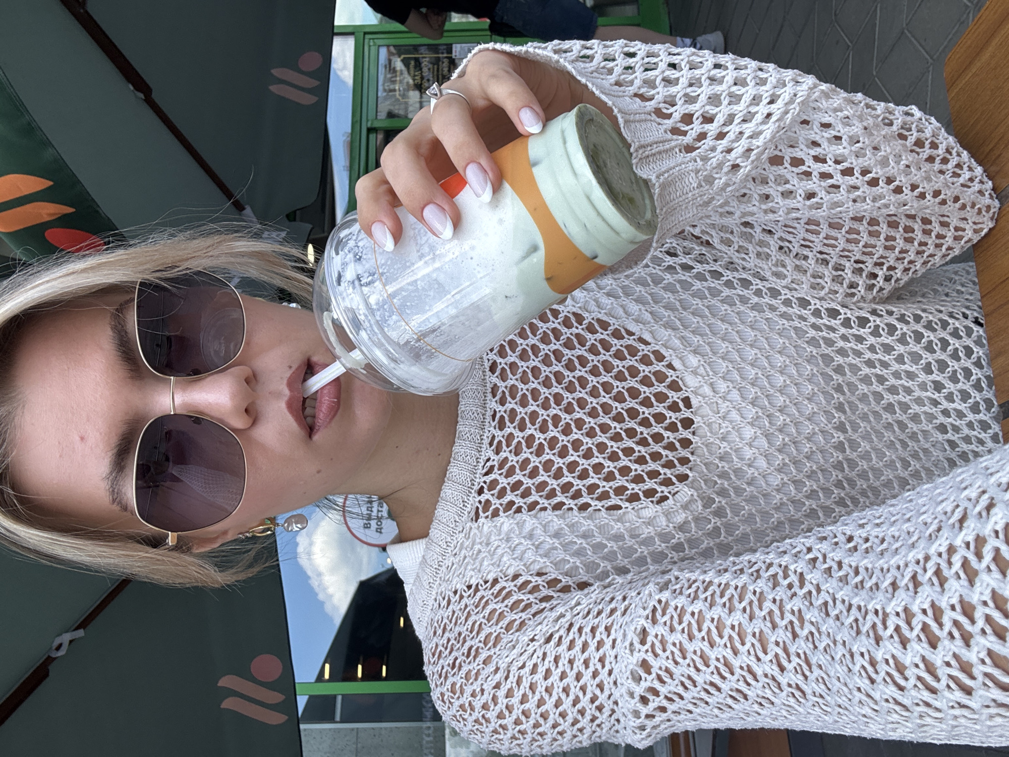 A woman wearing sunglasses and a white crocheted sweater is drinking from a nearly empty plastic cup with a straw. The cup has a green lid and an orange band. She is seated at a table outdoors, under an umbrella, near a building with green accents and the words "ВЫДАЧА ДОСТАВКИ" on a sign. The location is in the city of Unknown, Unknown.