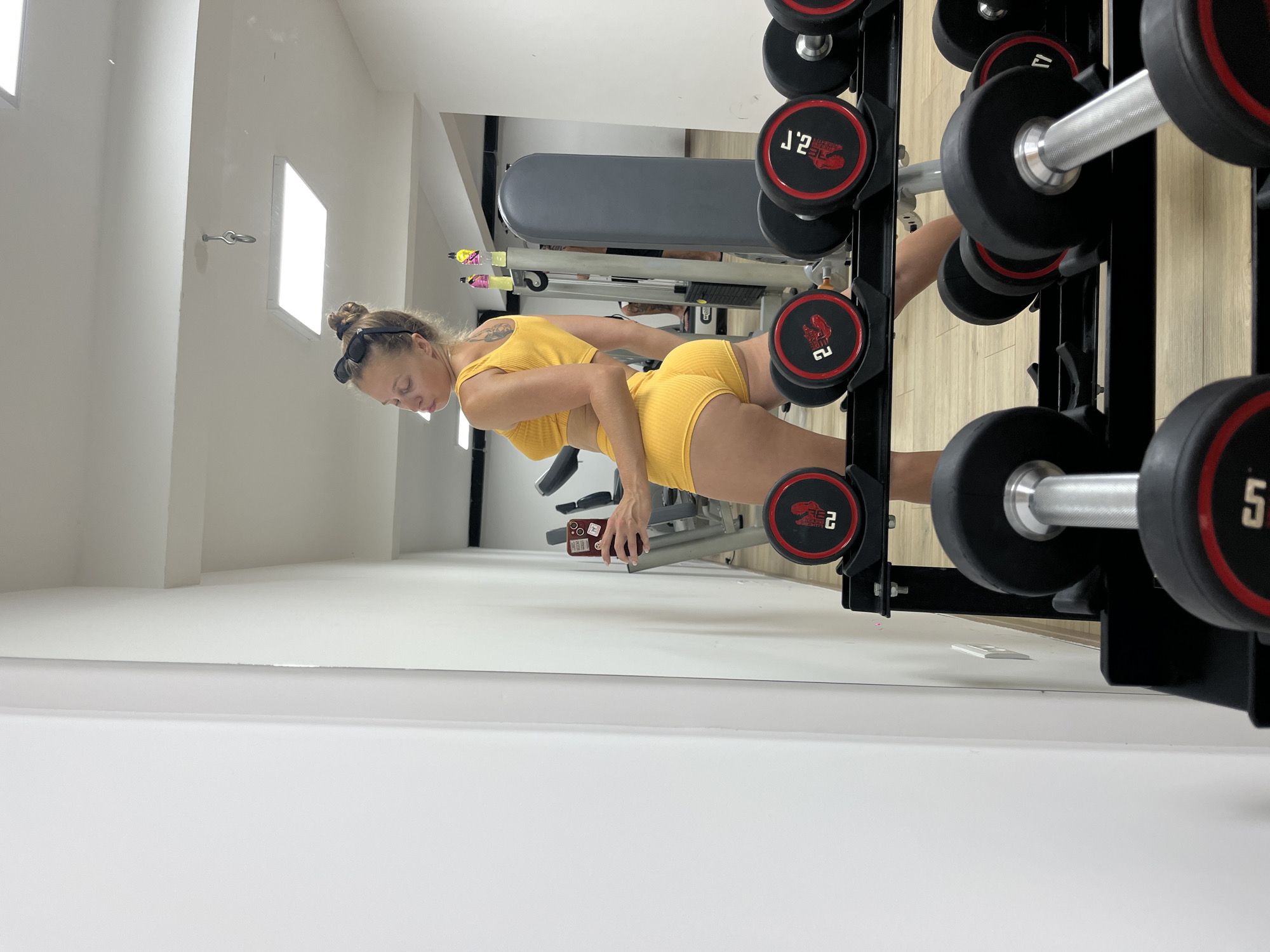 A woman in a yellow athletic outfit stands in a gym and takes a selfie in a mirror. She is wearing a yellow sports bra and matching shorts. Her hair is pulled up in a bun, and she is wearing sunglasses on top of her head. In the foreground, a rack of dumbbells is visible. In the background, gym equipment, including a weight machine, can be seen. The walls of the gym are white, and the floor is made of light-colored wood.