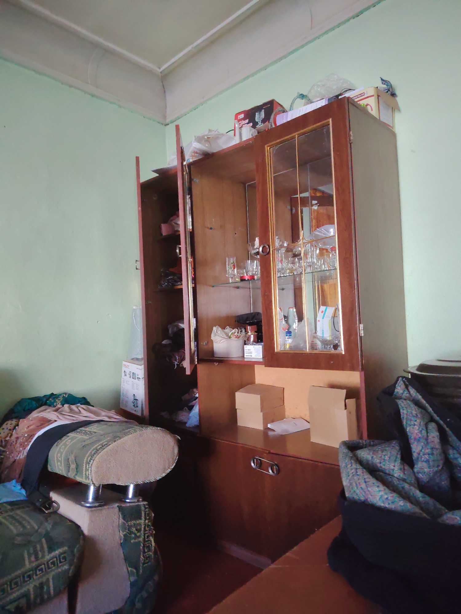 The image depicts the interior of a room, likely a living room or dining room, with light green walls and a white ceiling. A large wooden cabinet with glass doors is the central focus. This cabinet is filled with glassware, some boxed items, and various other objects. To the left of the cabinet, a portion of a sofa or seating area is visible, covered with fabric and a black strap. To the right of the cabinet, a dark-colored pile of fabric or clothing is present. Several cardboard boxes are stacked on a lower shelf of the cabinet. The scene suggests a cluttered yet lived-in space. The location in Almachi, Uzbekistan, is not directly evident from the visual cues within the image.