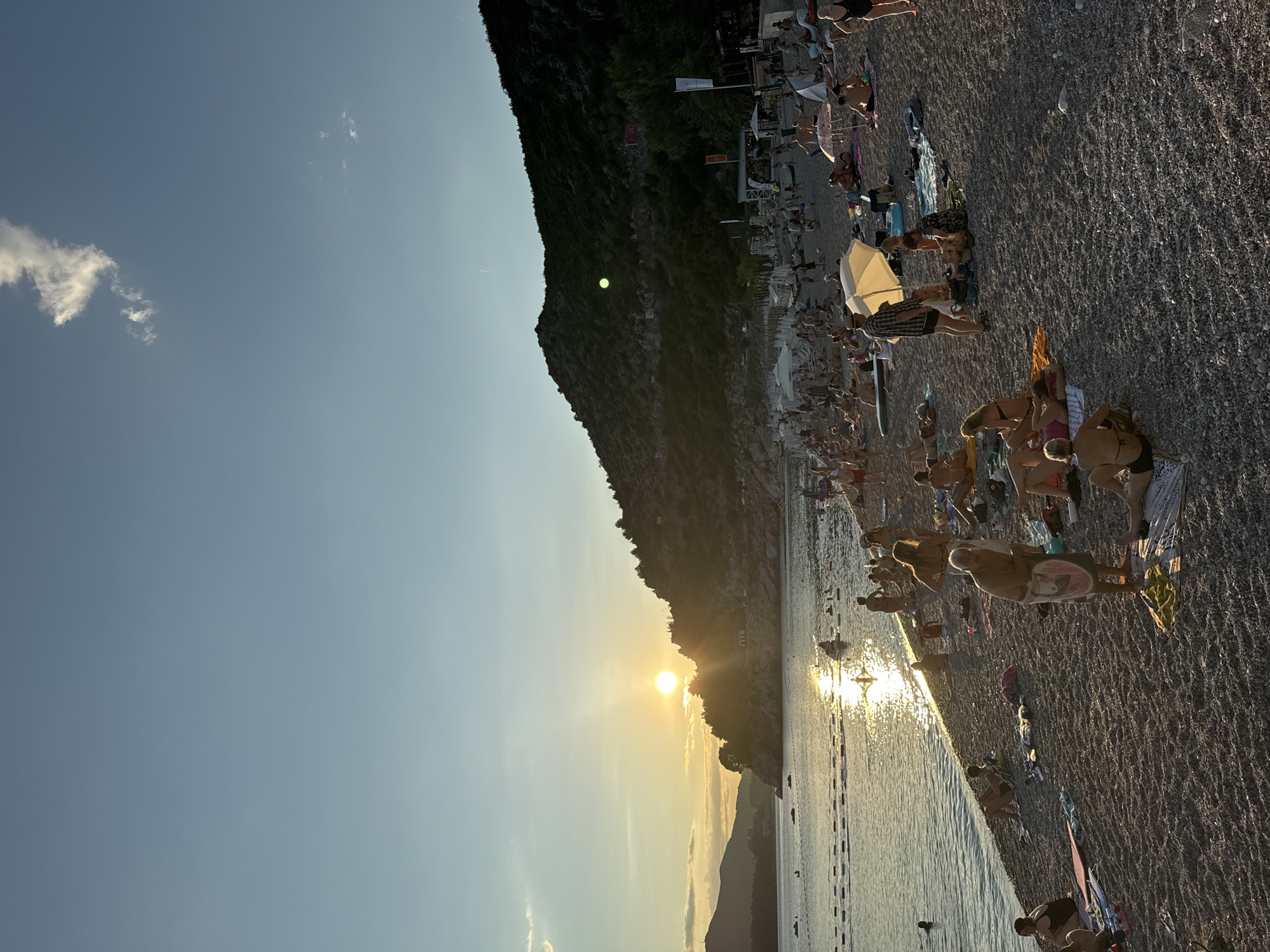 This image captures a sunset scene at Kamenovo beach in Montenegro. The sun, low in the sky, casts a warm glow across the water, creating a shimmering reflection on the sea's surface. The beach is populated with numerous individuals, many of whom are sunbathing or relaxing on towels and mats spread across the pebble shore. Some are swimming or wading in the shallow water, while others appear to be socializing.
The background features a rocky, tree-covered hillside that slopes down towards the sea, providing a dramatic natural backdrop to the bustling beach. A few structures, possibly beachside amenities or small buildings, are visible nestled into the hillside. The sky is a clear blue with a few wispy clouds, indicating fair weather. The overall atmosphere is one of leisure and recreation, with people enjoying the late afternoon sun and the coastal environment.
