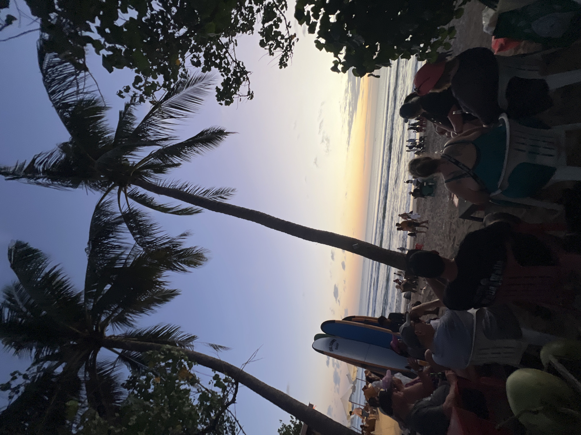 The image depicts a tranquil beach scene in Kuta, Indonesia, during twilight. Two prominent palm trees frame the shot, their fronds silhouetted against a gradient sky of deep blue fading into a soft orange and yellow sunset. In the foreground, a group of people are seated on the sand, facing the ocean. Some are seated in chairs, while others are on the ground. Two surfboards are propped up near the group, suggesting a connection to surfing activities. The ocean is calm with gentle waves rolling onto the shore, and a few distant figures can be seen walking along the beach. The atmosphere appears relaxed and communal, with people gathered to enjoy the fading light and the ocean breeze.