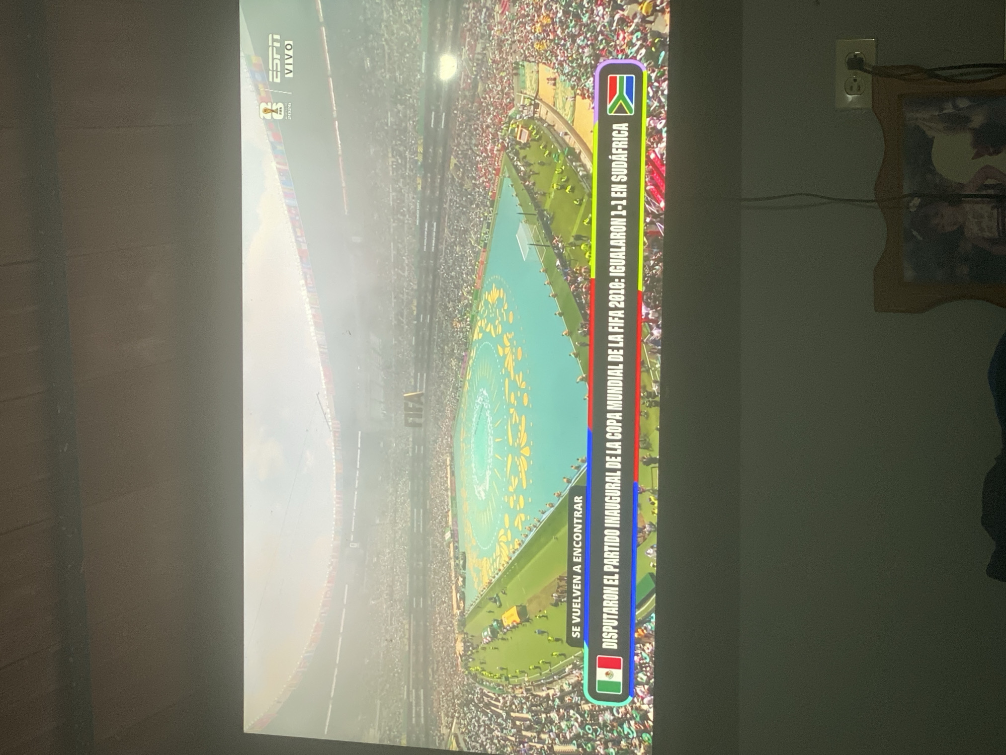 The image shows a television screen displaying a soccer match, possibly from the FIFA World Cup. The screen shows a packed stadium with a green field and yellow patterns. Along the bottom of the screen, there is a banner with text in Spanish and flags. The text appears to read "SE VUELVEN A ENCONTRAR DISPUTARON EL PARTIDO INAUGURAL DE LA COPA MUNDIAL DE LA FIFA 2010: IGUALARON 1-1 EN SUDAFRICA" which translates to "They meet again They played the opening match of the FIFA World Cup 2010: they drew 1-1 in South Africa." The flags displayed are the Mexican flag and the South African flag. There are also logos for ESPN and FIFA. The scene is viewed from an upward angle, suggesting the television is mounted or placed at a height. The lower portion of the image shows a wall with a power outlet and a framed picture. There is no indication of the city of El Cambur, Venezuela within the provided image.