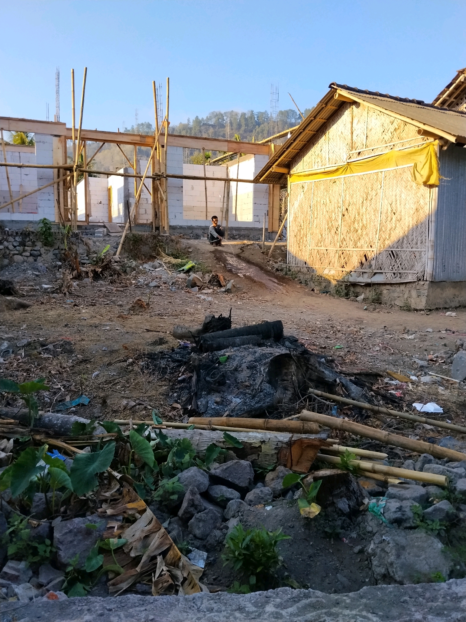 The image depicts an outdoor scene in what appears to be a rural or semi-urban area, possibly Tlogosari, Indonesia, given the contextual information. The foreground is cluttered with rocks, debris, and dried plant matter, including a pile of burnt logs and branches. In the middle ground, there is a construction site with walls made of white bricks and bamboo scaffolding. A young person is sitting on a dirt mound near the construction. To the right, a small building constructed with woven bamboo walls and a corrugated metal roof is visible. The background shows a hillside with vegetation and some distant structures. The lighting suggests it is either early morning or late afternoon due to the long shadows. The overall impression is one of ongoing construction in a somewhat unkempt environment.
