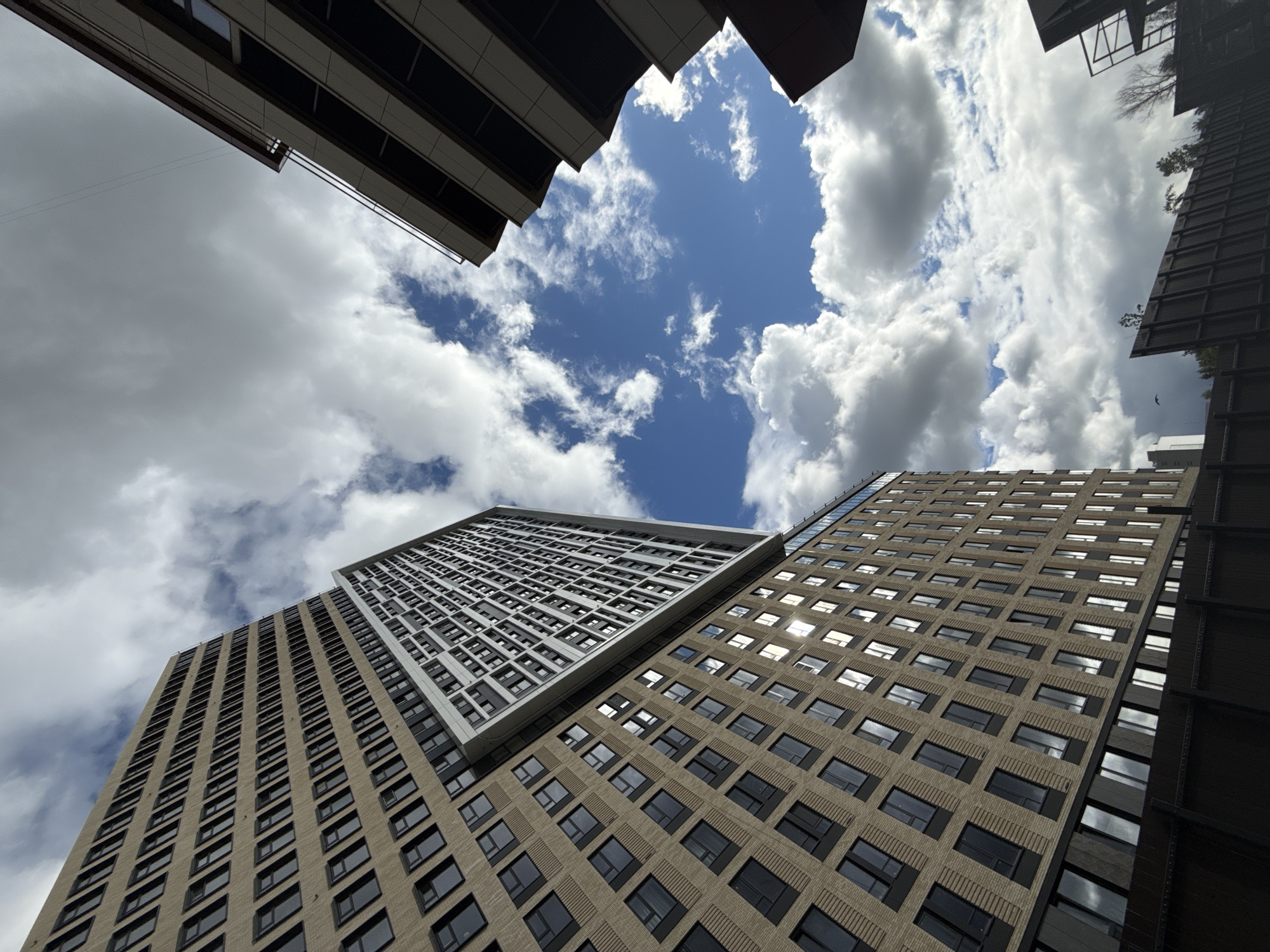 The image is a low-angle shot of several tall buildings in Khimki, Russia, viewed from below, creating a perspective that emphasizes their height. The buildings are modern, with a grid-like pattern of windows covering their facades. One building has a light-colored, almost white exterior, while the others have a beige or light brown color. The sky is visible between the buildings, featuring a mix of blue patches and white, fluffy clouds. The composition is dominated by the architectural forms and the interplay of light and shadow on the buildings and clouds.