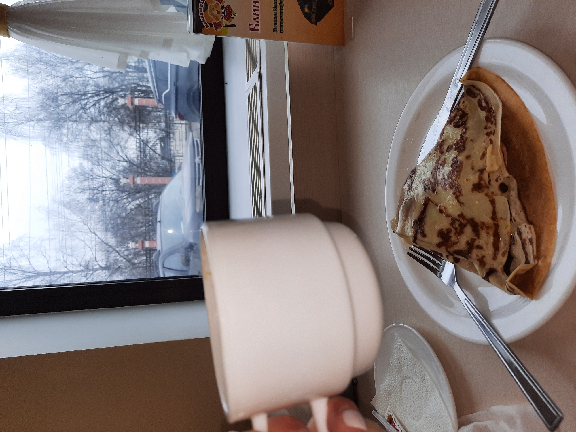 A top-down shot captures a breakfast scene indoors, near a window. A white plate holding a folded pancake with dark drizzles, and cutlery (knife and fork) lie on a neutral-toned surface. Beside it, a small plate with napkins sits partially obscured by a white mug being held. In the background, a window displays a view of bare trees, a gray sky, and vehicles, suggesting a cold or winter setting. The presence of a "Blini" (Pancake) package indicates a breakfast setting, possibly in a cafe or restaurant. The location is Izhevsk, Russia, as indicated by the Russian language on the "Blini" package, and the general scenery aligns with locations in that region. There is a partial view of someone's hand holding the mug. The scene is undisturbed, and the only actions implied are eating and drinking.