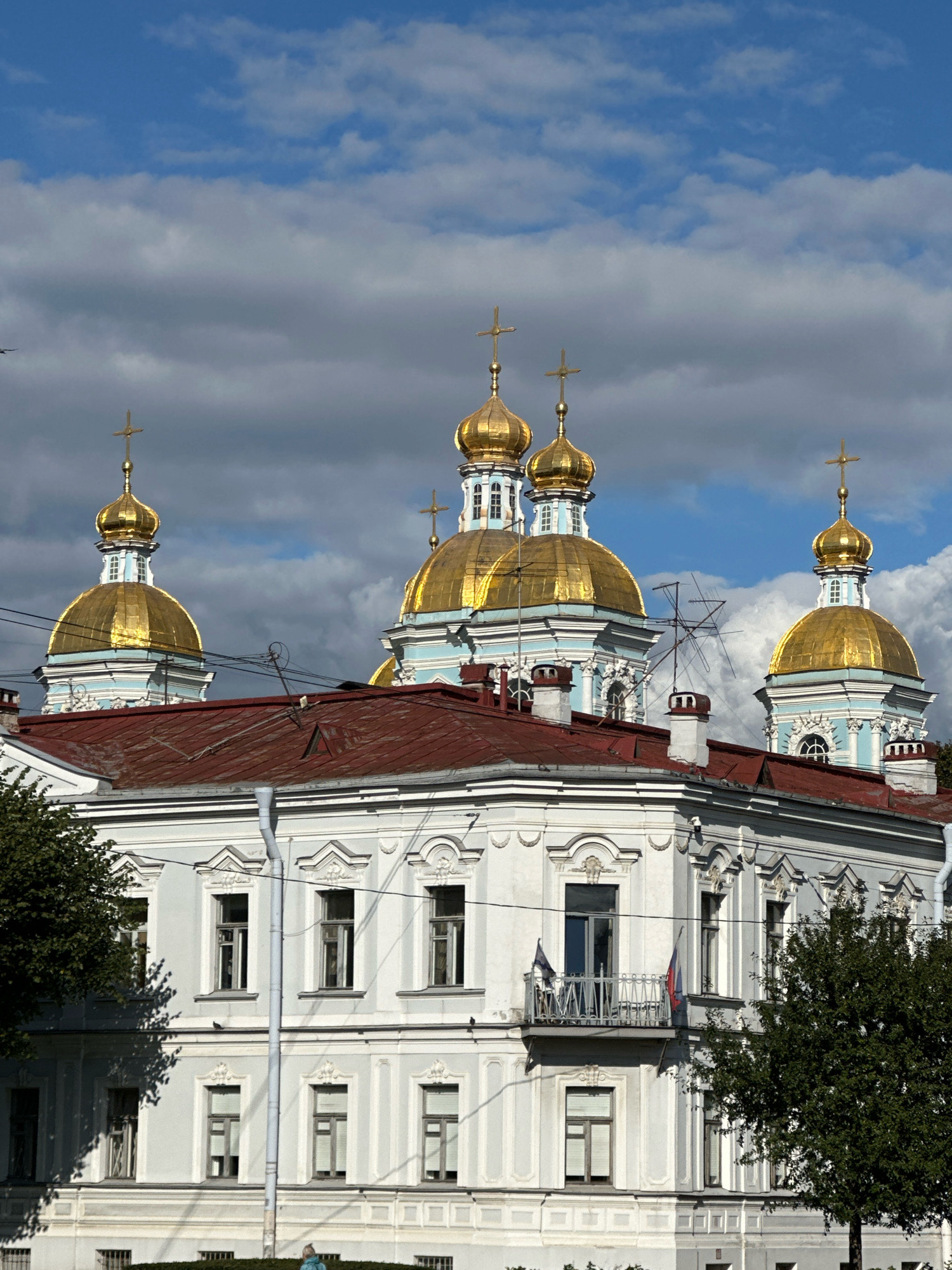 This is a vertical, medium shot of the Saint Nicholas Naval Cathedral in St. Petersburg, Russia, on a partly cloudy day. The bright, gilded domes of the cathedral dominate the upper portion of the frame, set against a bright blue sky with scattered white and grey clouds. The main body of the cathedral, with its pale blue walls and white architectural details, is visible behind the red-tiled roof of a foreground building. This foreground building is light grey with white decorative elements around the windows and cornices. Several windows are visible, some with their shutters partially closed. A balcony with a decorative railing and two flags, one partially visible, projects from the building's facade. Trees with dark green foliage flank the foreground building on both sides. Electrical wires crisscross the scene, and a few small birds are visible in the sky.