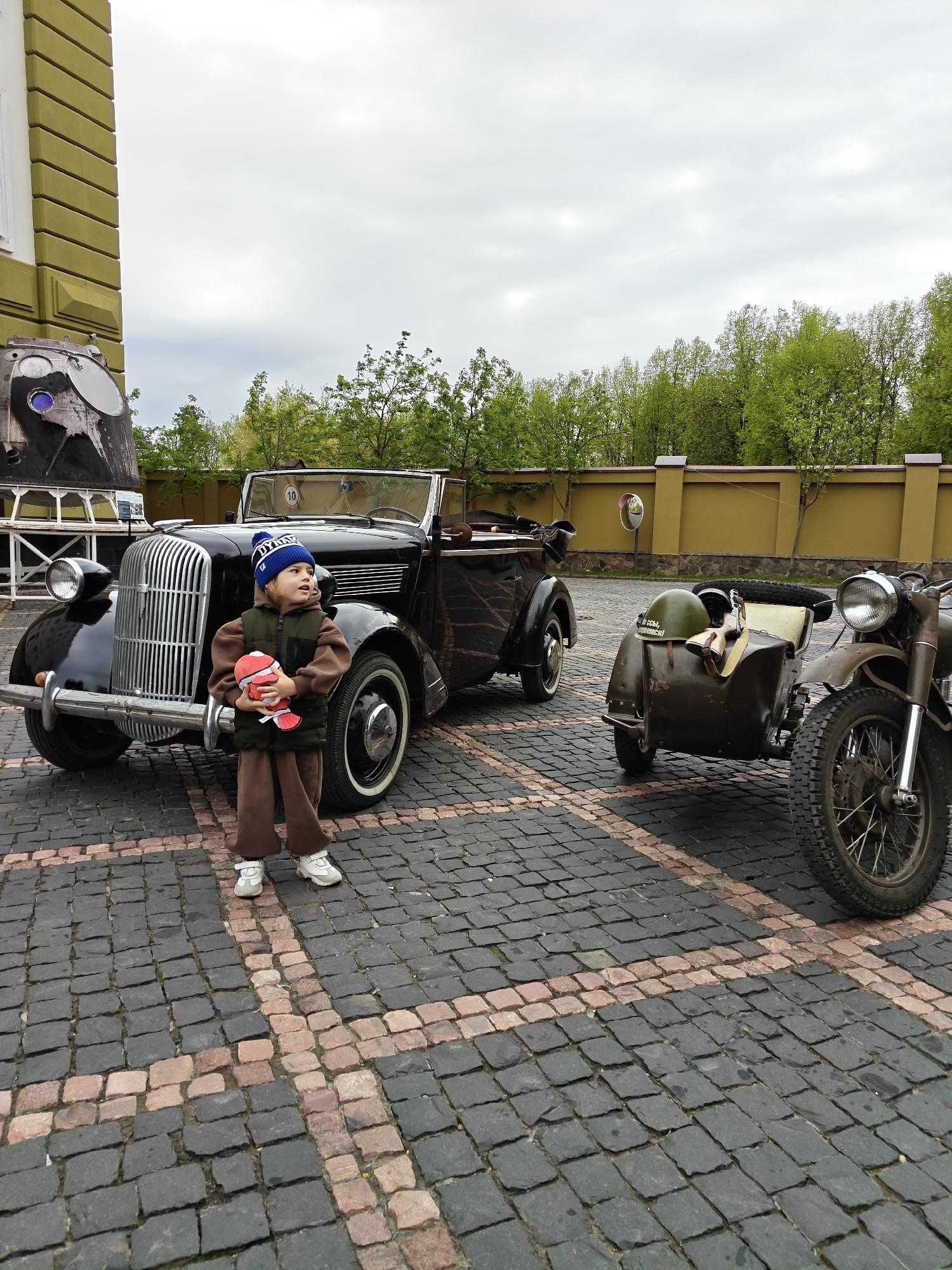 A young child, wearing a blue hat, brown jacket and pants, and white sneakers, stands in front of a black vintage car, holding a red stuffed animal. To the right of the car is a motorcycle with a sidecar. The scene is set in Архангельское (красногорский Район), Russia, evident by the architecture and vehicles. The ground is cobblestone. There is a building to the left and a wall with trees behind it in the background. The sky is overcast.