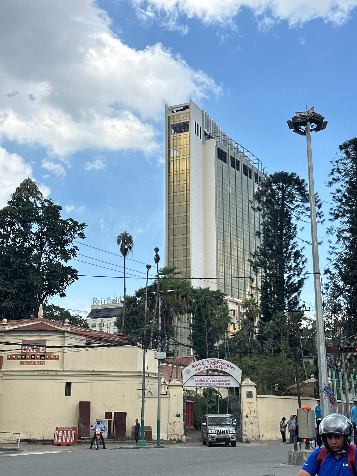 A wide shot captures an urban scene in Kathmandu Metropolitan City, Nepal, under a partly cloudy sky. In the background, a modern, tall building with a golden facade dominates the skyline. In the mid-ground, a lower, cream-colored building with "CAFE RESTAURANT" signage is visible. Adjacent to it, an arched gateway with "KAISER LIBRARY" and "Public Procurement Monitoring Office" inscribed on it marks an entrance. Several vehicles, including a silver SUV and a motorcycle with a rider wearing a helmet, are present on the paved road in the foreground. Pedestrians are seen near the gateway. The scene is dotted with lush green trees and utility poles.