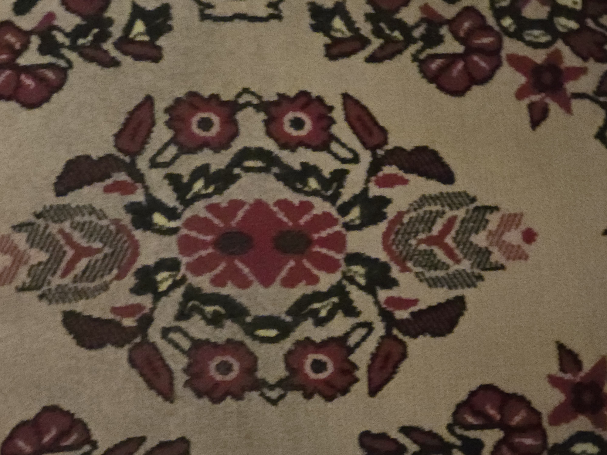 The image shows a close-up, detailed view of a carpet with a floral pattern. The carpet is predominantly a light beige color, serving as the background for the intricate design. The pattern features a central, large, multi-layered flower in shades of deep red and dark red, with a dark center. Surrounding this central element are smaller floral motifs, also in reds and dark reds, interspersed with dark green leaves and some lighter yellow accents. The leaves are stylized and pointed. There are also geometric or tribal-like motifs in dark gray, red, and beige that appear above and below the central floral arrangement. The overall impression is of a traditional, possibly handmade, textile. The lighting appears to be uniform, highlighting the texture and weave of the carpet.