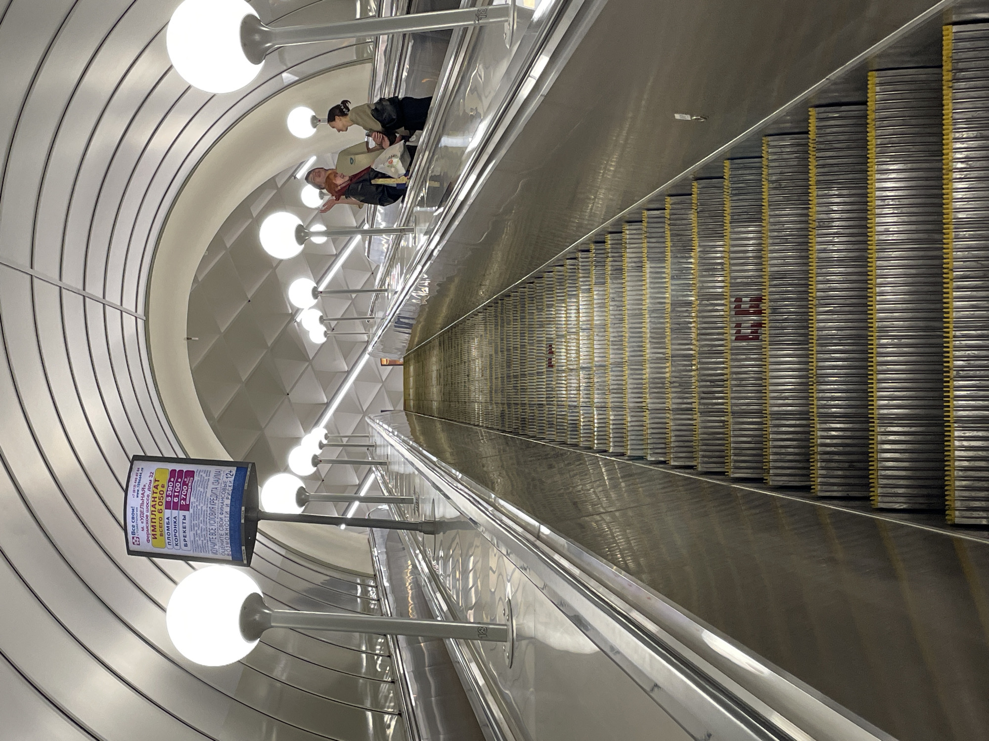 The image captures the interior of a subway station in Saint Petersburg, Russia. It shows a long escalator descending into the station. The escalator is lined with silver steps, and yellow safety guides.
Several globe-shaped lights illuminate the scene, attached to posts along the escalator. The station's walls and ceiling are predominantly white. An advertisement for dental implants is displayed near one of the lights.
Two people can be seen on the escalator. One is a redheaded person, and the other person has their hair in a bun.