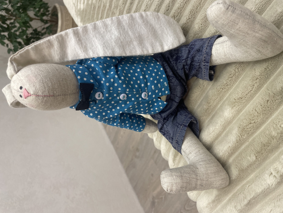 A handmade plush rabbit doll is depicted seated in an indoor setting in Izhevsk, Russia. The doll is constructed from light beige fabric, featuring long, floppy ears, stitched black eyes, a pink triangular nose, and a single pink line for a mouth. It wears a teal blue long-sleeved shirt with white polka dots, a dark navy blue bow tie, and dark blue patterned denim-like shorts. The doll is positioned sitting on a ribbed, light cream-colored plush surface. In the background, a light-colored textured wall is visible on the left, and a portion of a green potted plant in a light-colored pot is discernible on the right.
