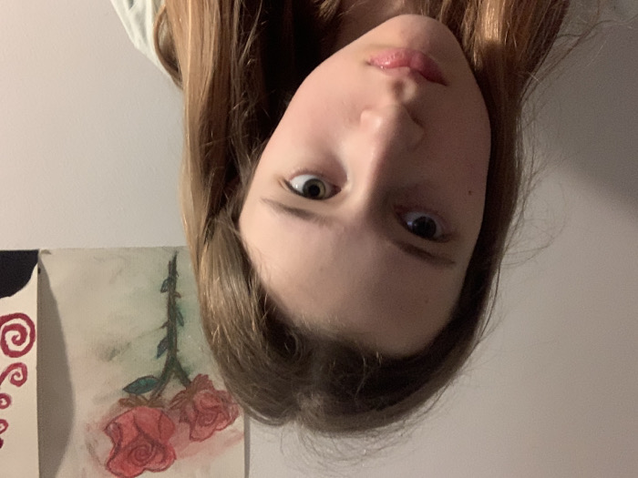 The image features a close-up, selfie-style portrait of a young individual, likely female, captured from an overhead or upward-looking perspective. The original image is rotated 90 degrees clockwise; when viewed upright, the person's face occupies a significant portion of the lower and central frame, looking up towards the camera.
The individual has light to medium brown hair that appears long and falls around their face. Their eyes are open and directed towards the viewer, with visible dark pupils and irises that seem to be a dark brown or hazel color. Their lips are slightly parted, showing a natural, glossy pink hue. The facial expression appears neutral or slightly curious.
In the background, to the left of the person's head (when viewed upright), there is a plain, light-colored wall. Below and to the left of the person (when viewed upright), two pieces of artwork or canvases are visible, suggesting an indoor setting like a room or studio.
The artwork on the right, closer to the person, depicts two red roses with green stems and leaves, rendered in a style that resembles watercolor or colored pencil. The stems appear slightly thorny. The artwork on the left, partially obscured, features a design with red swirls or spirals on a light background, with a dark, possibly black, element at its upper left edge.
The lighting in the image is soft and even, suggesting indoor artificial light or diffuse natural light. There is no visible text in the image.