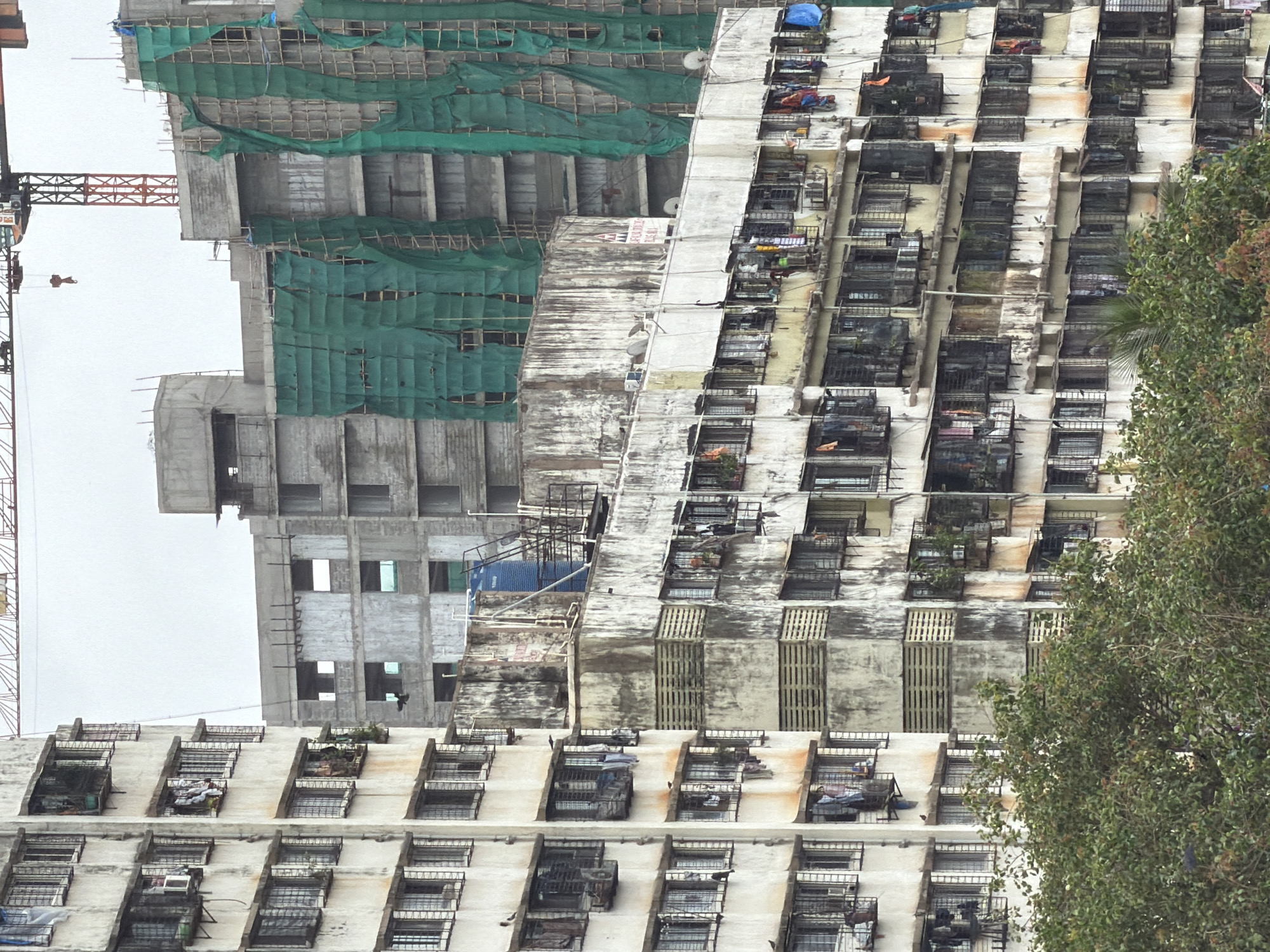 The image shows a dense urban landscape in Mumbai, India, focusing on a cluster of multi-story residential buildings and a partially constructed high-rise. The residential buildings, likely older, exhibit signs of wear and tear with stained facades, exposed pipes, and numerous balconies adorned with clothing, satellite dishes, and air conditioning units. Many windows are covered with protective bars or grills. Construction is underway on a larger building in the background, draped in green netting, with a prominent orange construction crane visible against a hazy sky. The scene is typical of Mumbai's vertical expansion, where older housing coexists with ongoing development.
