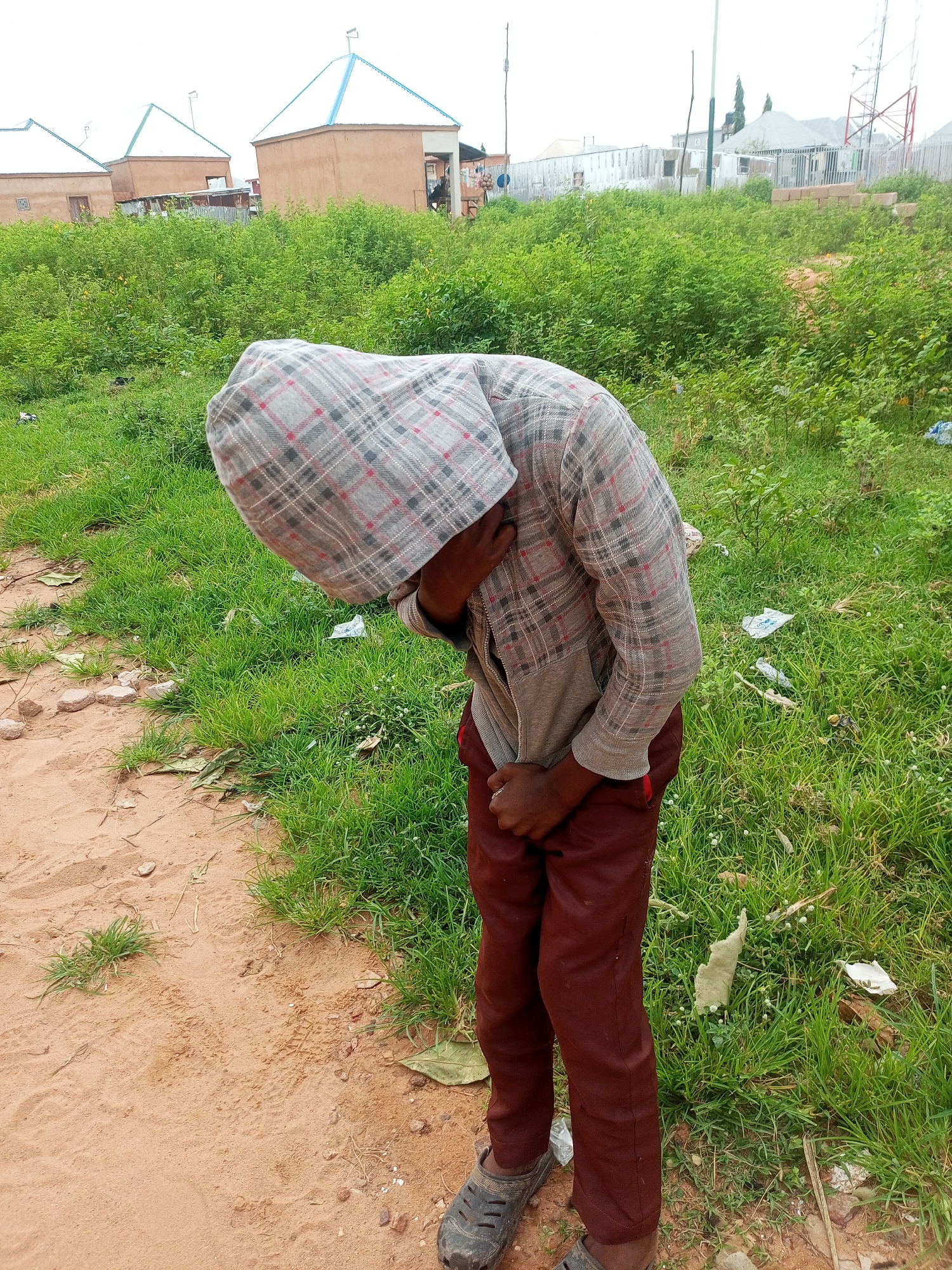 A young person wearing a plaid hooded jacket and maroon pants is standing outdoors in Gombe, Nigeria, with their head bowed and hands in their pants. The person is on a dirt path next to a grassy area. In the background, there are several buildings with distinctive conical roofs and a telecommunications tower. The environment appears somewhat undeveloped, with scattered debris in the grass. The sky is overcast.