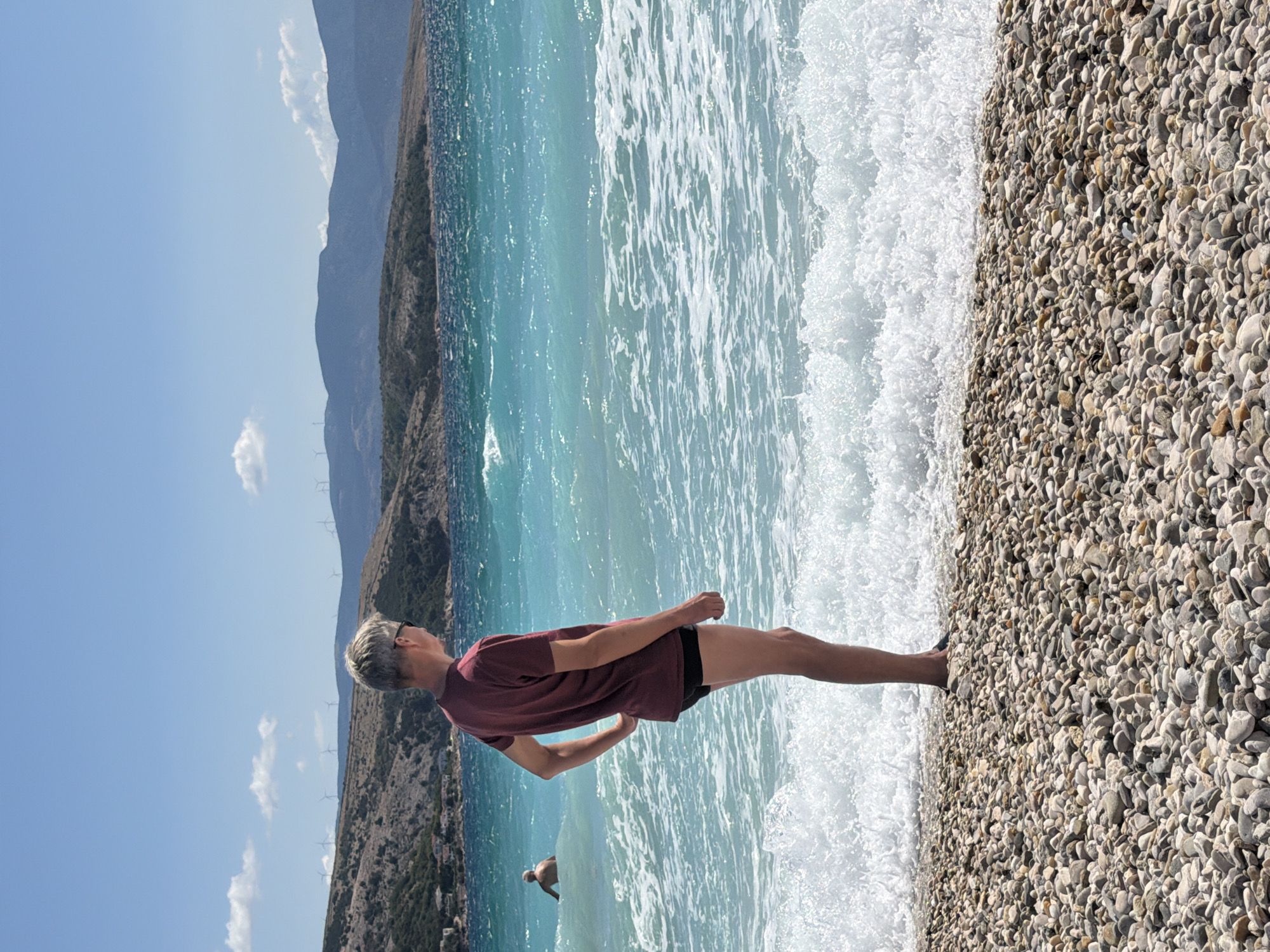 A middle-aged man with short, grey hair stands on a pebble beach, facing away from the camera, looking out at the turquoise sea. He is wearing a maroon t-shirt and black swim trunks. His right hand rests on his hip. In the background, the sea is choppy with white waves breaking on the shore. Further out, another person is visible partially submerged in the water. The coastline in the distance is mountainous and sparsely vegetated, with a few buildings scattered on a hillside. Wind turbines are visible on the crest of the hills. The sky is a clear blue with a few scattered white clouds. The scene is likely the town of Baška on the island of Krk in Općina Baška, Croatia, a popular tourist destination known for its beaches and scenic landscapes.