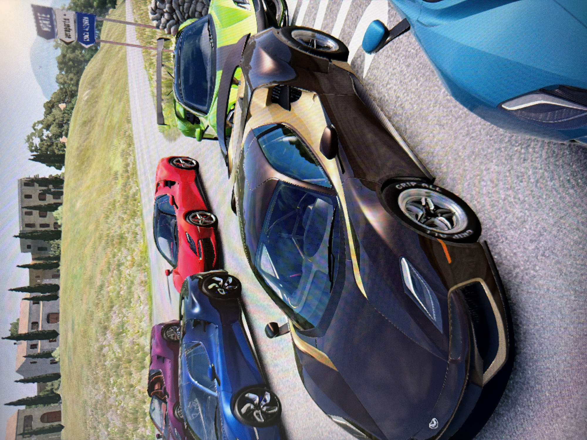 A vibrant screenshot from a video game shows a lineup of several high-performance sports cars parked on an asphalt road in a scenic, mountainous countryside setting during daytime. The image is taken from a slightly elevated, side-front perspective, looking down the row of vehicles.
In the immediate foreground, a sleek, dark grey or black supercar with prominent gold accents and dark tinted windows dominates the frame. Its front right wheel is visible, with "GRIP ALL" clearly printed on the tire. Partially visible to the right of this car is the front quarter of a bright blue car. Behind the dark grey car, a lime green supercar is parked, followed by a bright red car, then a dark blue car, and finally a purple car further down the line. All cars exhibit futuristic and aerodynamic designs.
The road surface is a textured grey asphalt, bordered by a grassy shoulder. In the background, rolling green hills dotted with trees rise towards distant, hazy mountains. On the left side of the road, several Mediterranean-style buildings, possibly houses or villas, are visible, interspersed with tall, narrow trees.
Two road signs are present in the mid-ground: a blue sign with "AIRPORTS" clearly legible at the top, and another word below it that is indistinct. Next to it, a white sign shows a word that appears to start with "VILLA" or "VALLE" followed by "TE" or "TÈ". The sky is bright and clear, indicating sunny weather. The overall impression is that of a grand event or a starting grid in a racing game.