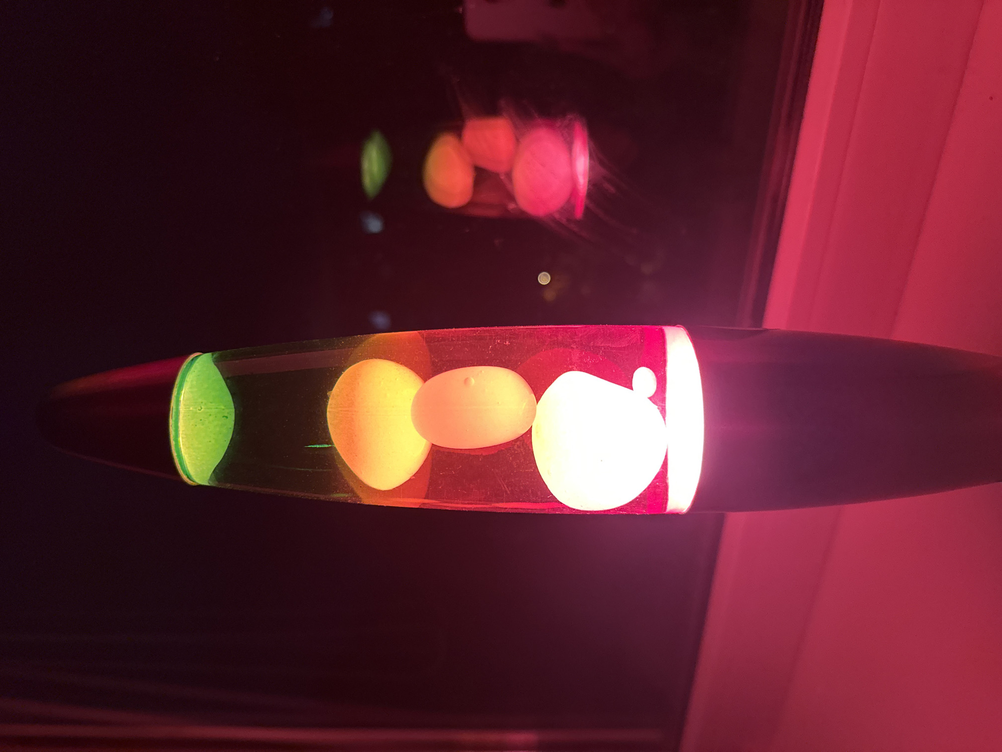 The image shows a lava lamp in operation, displaying a sequence of colored blobs. From left to right, there is a green blob, followed by yellow, then a light orange and finally a white blob. Reflections of these blobs are visible on a dark background above the lamp. The lamp itself has a dark metallic structure, and it is positioned near a light red colored vertical surface, possibly a wall. The location is unknown.