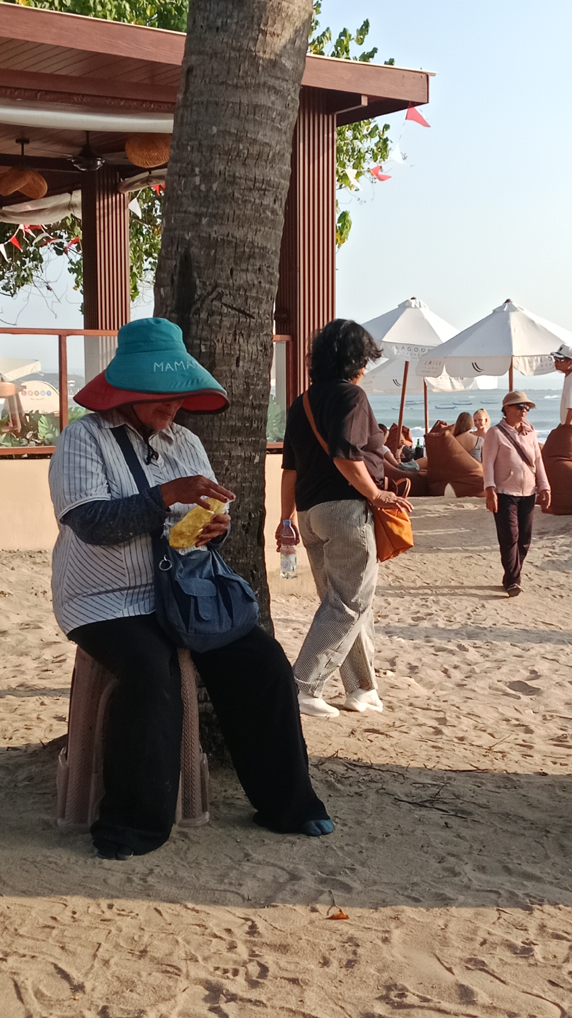 A medium shot shows a woman seated on a stool on a sandy beach in Kuta, Indonesia. She is wearing a large, multi-colored hat, a striped shirt, and black pants. She is holding a yellow object, possibly food, in her hands and appears to be eating it. A blue bag is slung over her shoulder. To her right, another woman walks away from the camera, wearing striped pants and a black shirt, with an orange bag over her shoulder. In the background, beach umbrellas and beanbag chairs are visible, suggesting a resort or public beach area. The sun is shining, casting shadows on the sand, and the sky is clear and blue. A palm tree trunk is prominently positioned to the left of the seated woman. The scene suggests a relaxed atmosphere on a tropical beach.
