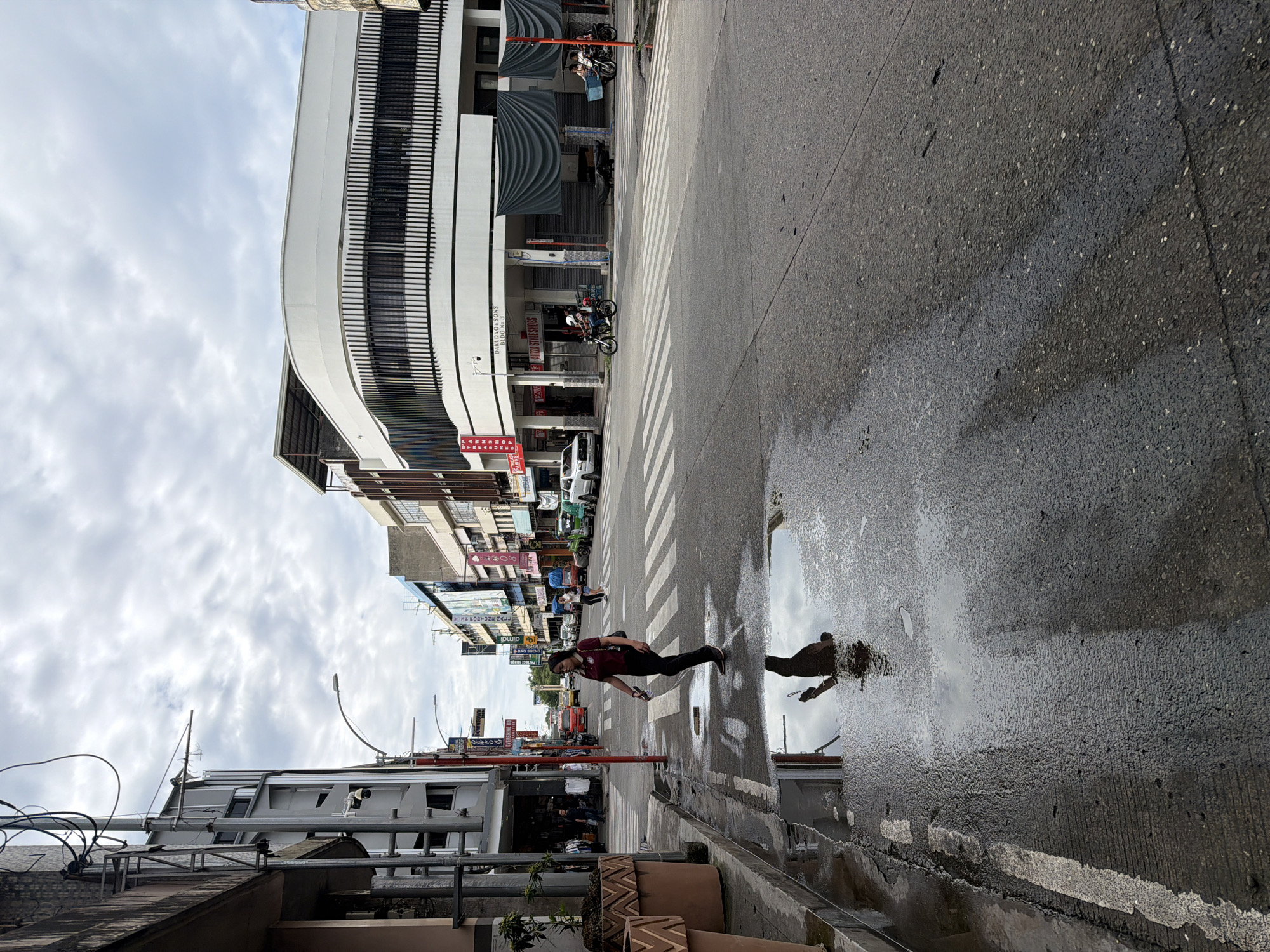The image depicts a street scene in Davao City, Philippines. A person wearing a maroon shirt and black pants is walking across a pedestrian crossing. A puddle on the street reflects the person and the surrounding buildings. Buildings line both sides of the street with various signs and architectural styles. Motorbikes are parked near the crosswalk. The sky is overcast.