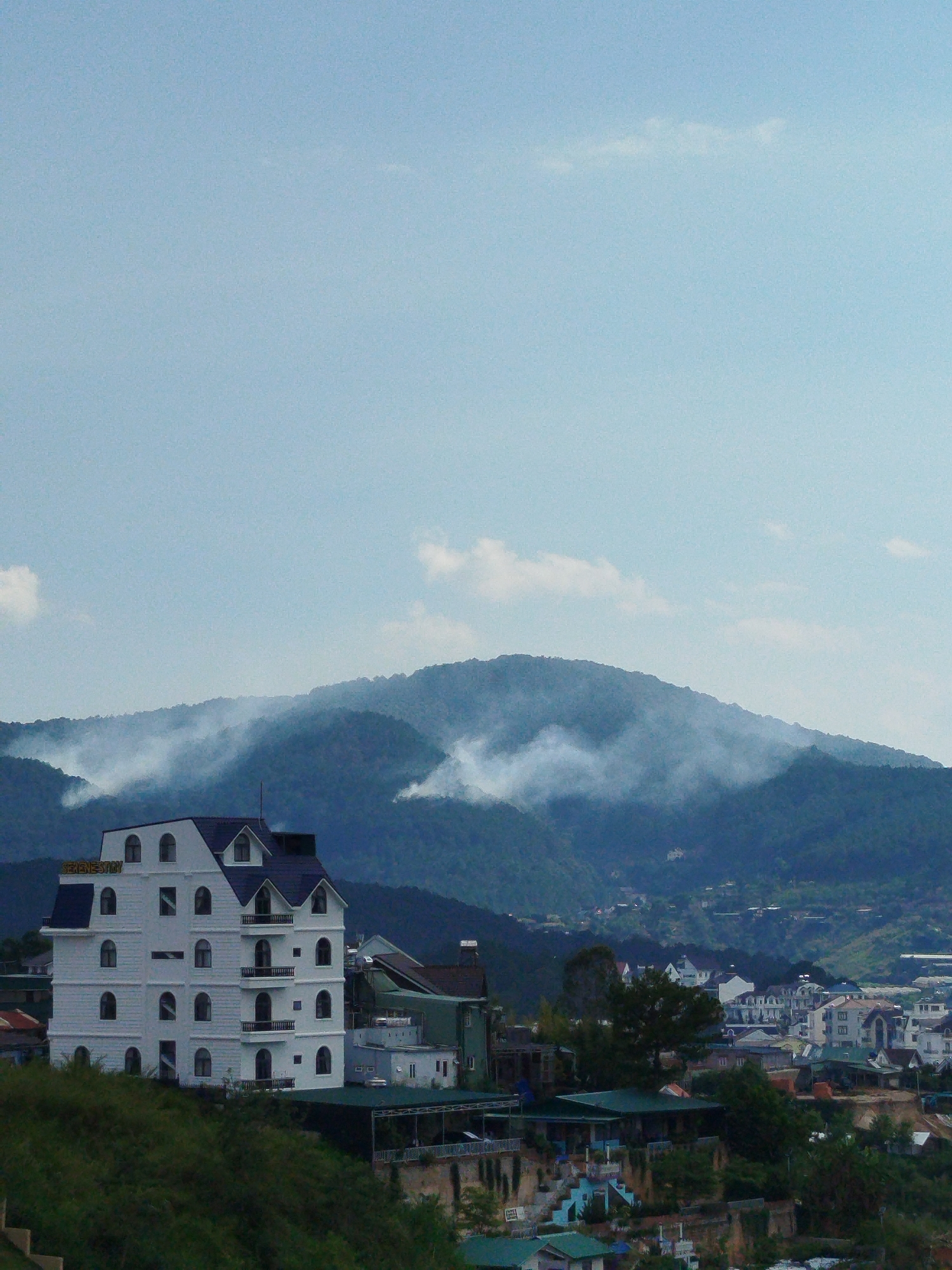 A view in Da Lat, Vietnam presents a tall, five-story, white building with a dark blue roof, located on an elevated green mound. The building has a sign reading "SERENESTRY" on its rooftop. Arched windows and small balconies are visible on the facade. Behind the building, a landscape of green hills and mountains is partially obscured by a layer of mist or low-hanging clouds. A cluster of other buildings and residences with varied architecture are seen scattered across the hillside. The sky above is a light blue with scattered clouds.