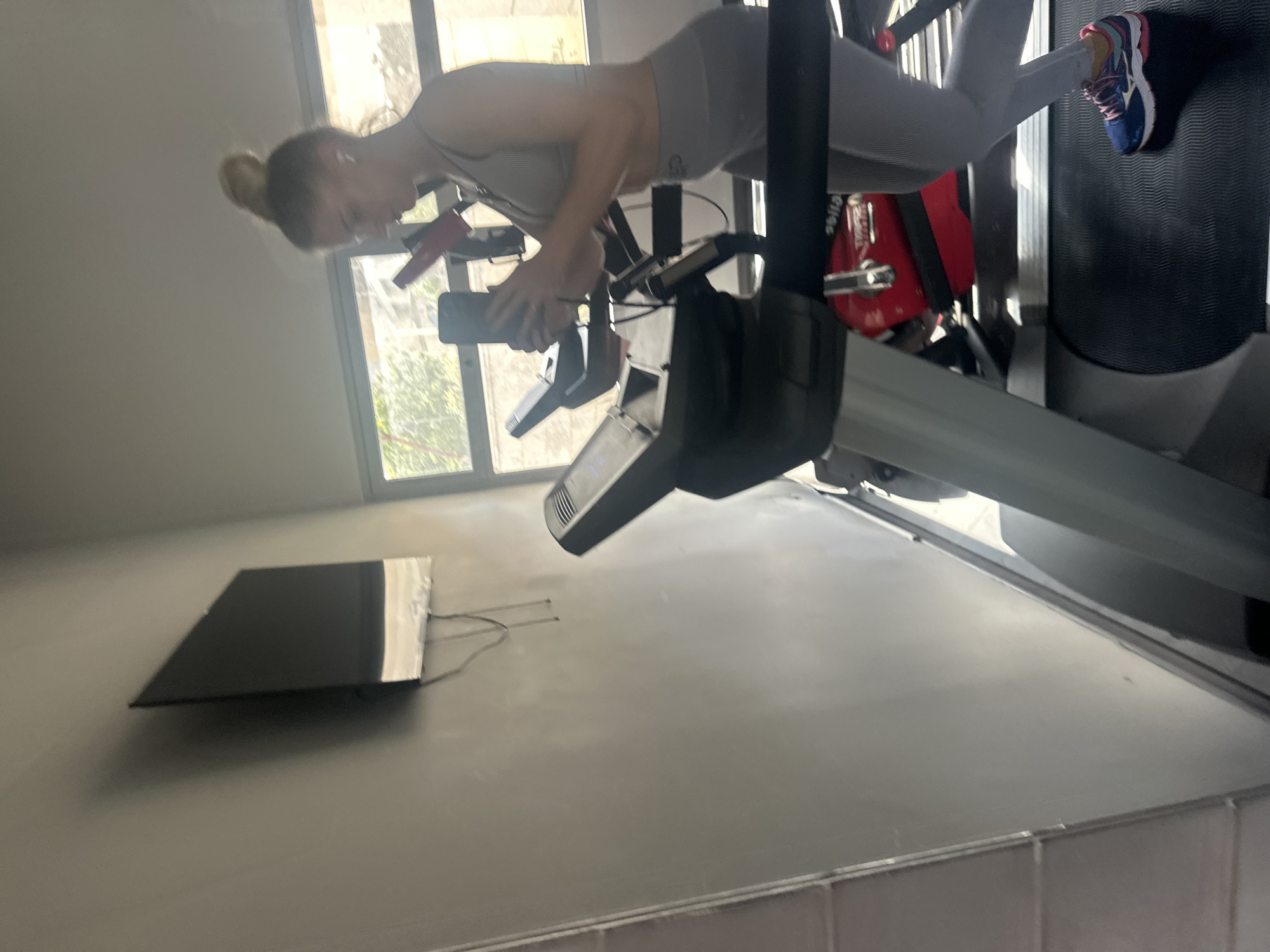 A woman in gray athletic wear and blue and pink sneakers is exercising on a treadmill in a room with gray walls. She is holding a smartphone and appears to be filming or taking a picture. There is a window behind her and a television mounted on the wall. The location is within the city of Petah Tikva, Israel.