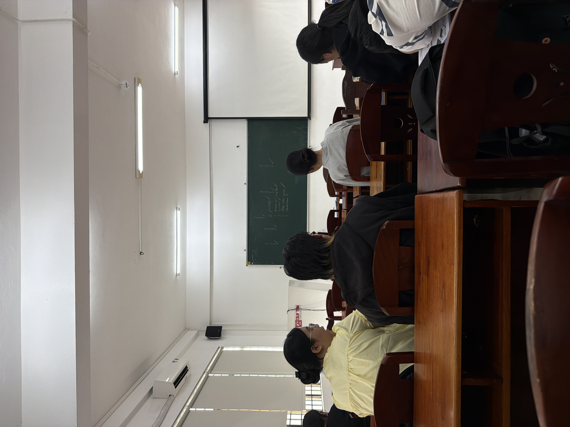 A classroom setting in Đà Nẵng, Vietnam, shows several students seated in rows of wooden desks, facing a blackboard and projector screen at the front. The room has white walls, fluorescent lighting, and an air conditioning unit. The students are engaged in learning.