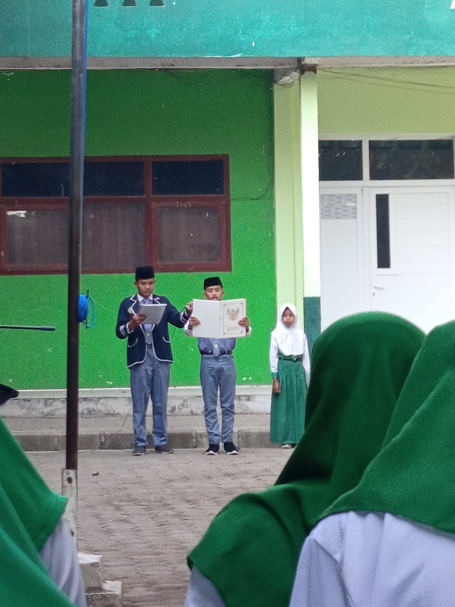 In Lumutan, Indonesia, two young males in formal attire stand on a paved area. The male on the left, wearing a dark blazer over a white shirt and vest, holds a tablet. The male on the right, in a similar outfit, holds an open book. Between them, a young female in a green skirt and white hijab stands facing forward. Behind the individuals is a green building with brown-framed windows and a light green accent wall. A white door with a dark vertical panel is visible to the right of the female. In the foreground, the heads and shoulders of three individuals wearing green hijabs are visible, obscuring the lower part of the scene. The ground is a dirt-like surface with some scattered debris. A dark grey pole stands to the left of the central figures.