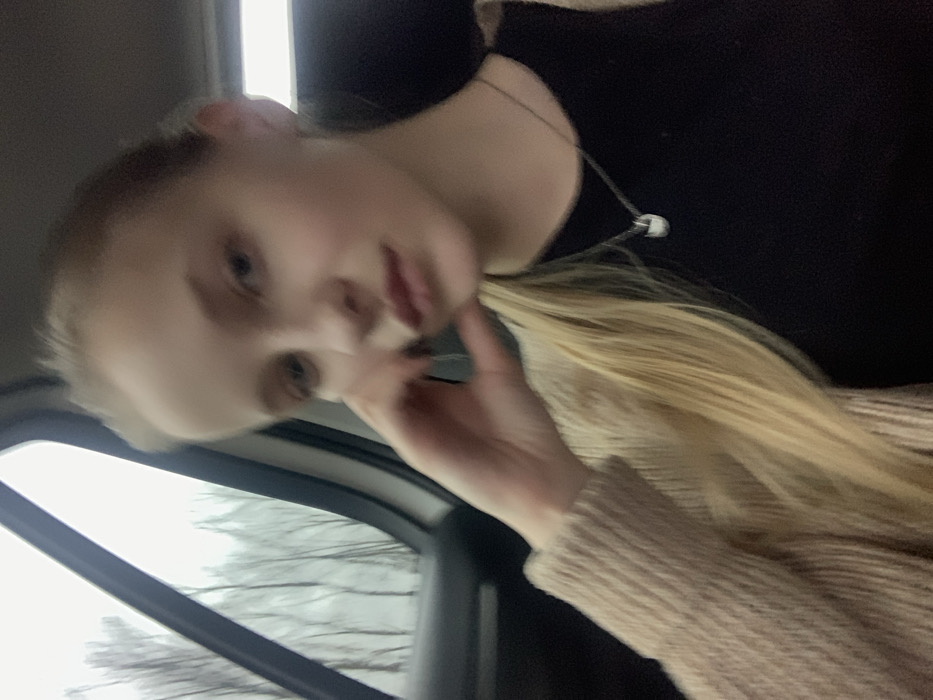 A young, fair-skinned female is depicted from the chest up, seated inside a vehicle. She has long, blonde hair, with a portion styled over her left shoulder. Her right hand is raised, with fingers resting against her chin and jawline. She is wearing a dark top, possibly black, underneath a light beige knitted cardigan with a visible ribbed texture on the sleeves. A silver-colored chain necklace with a small, rounded, light-colored pendant is visible on her chest. Her gaze is directed forward. Through the car window to her left, bare deciduous trees with thin trunks are visible against an overcast sky, suggesting a non-foliage season. The scene is located in Oliivka, Ukraine.