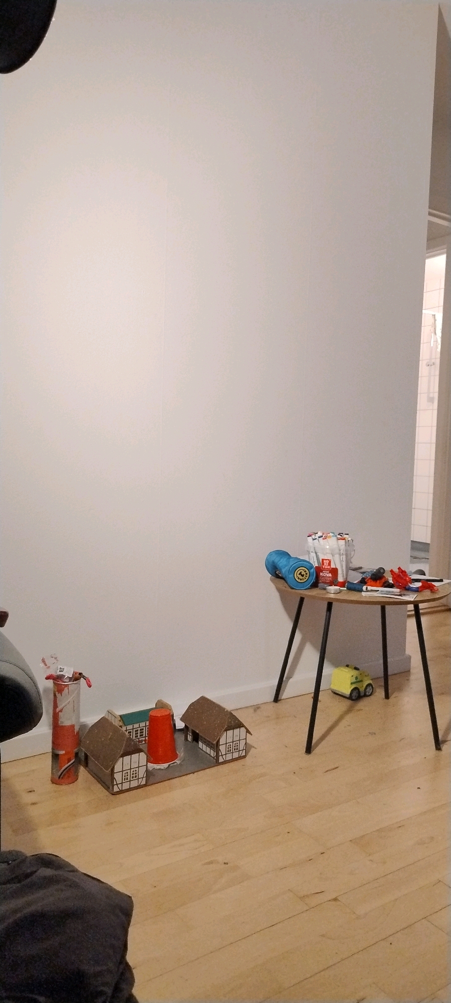 The image shows a domestic indoor scene, likely a child's play area, with elements suggesting a playful and creative environment. The setting is a room with light-colored walls and a wooden floor.
In the foreground, to the left, a dark gray, textured fabric object, possibly clothing or a cushion, is visible. To its right, a collection of miniature buildings, resembling houses with brown roofs and white-paneled walls, are arranged on a gray base. A bright orange conical object, possibly a cup or a toy, is placed between two of the houses. Next to the houses, on the far left, is an orange and white cylindrical object with some red on top, which appears to be a toy paint can or a similar item.
In the mid-ground, a small, round-topped table with three thin black legs stands on the wooden floor. On the table are various items, including a bright blue ball of yarn, a container of markers with colorful caps, a small red toy car or vehicle, and other scattered objects that are difficult to discern clearly. A small yellow toy vehicle, possibly a construction vehicle or a miniature ambulance, is positioned on the floor beneath the table.
In the background, on the right side of the image, a doorway or opening reveals a glimpse of another room, where white tiled walls and a shower curtain or door are visible, suggesting a bathroom or similar wet area. The overall impression is one of a child's play space, possibly in a residential area.
While the image provides visual details of a domestic interior, it does not contain any information that would allow for its specific location in Ishøj, Denmark, to be identified.