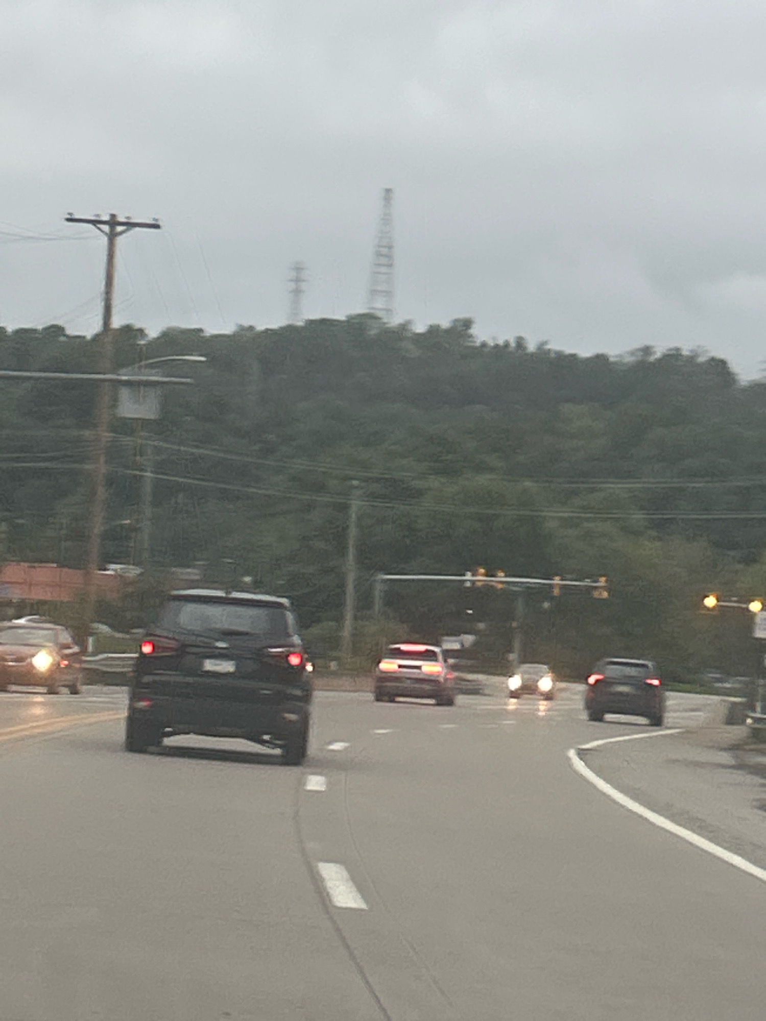 The image captures a view of a road in Carnegie, United States of America, on an overcast day. In the foreground, a black SUV is driving away from the camera. To the left, a red car is approaching, its headlights illuminated. Further down the road, other vehicles are visible, including a white SUV and another dark-colored vehicle. Traffic lights are present overhead, with some illuminated. The road is paved with white lane markings. In the background, a dense, green forest covers the hills. Utility poles with wires are also visible along the roadside, and a tall tower, possibly a radio or cell tower, stands on the horizon. The overall impression is a typical suburban scene on a cloudy day.