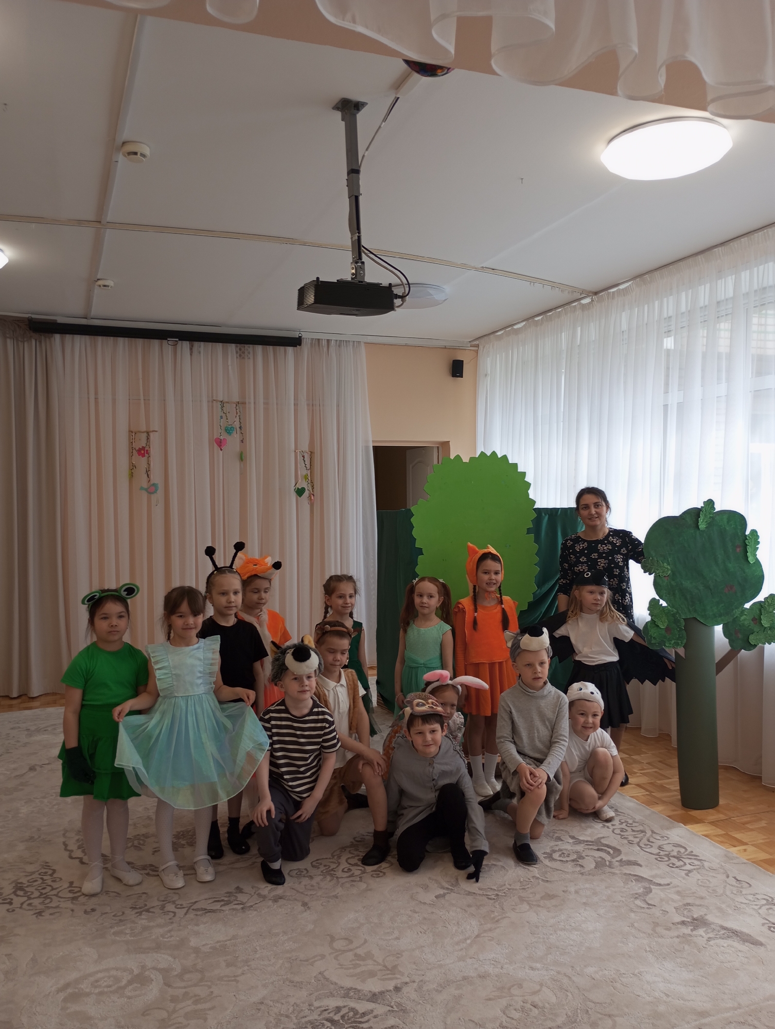 An indoor scene in Izhevsk, Russia, captures a group of approximately sixteen children and one adult woman posing for a photograph. The children, likely preschoolers or early elementary students, are dressed in various animal costumes, including a frog, a butterfly or ladybug (antennae), foxes, wolves, a hedgehog, rabbits, and a bat. They are arranged in several rows on a patterned rug, with some standing, some kneeling, and some crouching. A girl in a light blue dress is prominent in the front row. The adult woman, presumably a teacher or caretaker, stands smiling behind the children on the right side of the frame.
The setting appears to be a school or kindergarten hall, decorated with white sheer curtains covering large windows that allow natural light into the room. Two stylized green tree props are visible on the right, suggesting a backdrop for a play or performance. The ceiling features several light fixtures, including a large circular LED panel and a projector mounted near the center, along with two smoke detectors. A dark-colored speaker is mounted on the wall to the left. The overall atmosphere suggests a children's event, such as a school play or costume party. No visible text is present in the image.