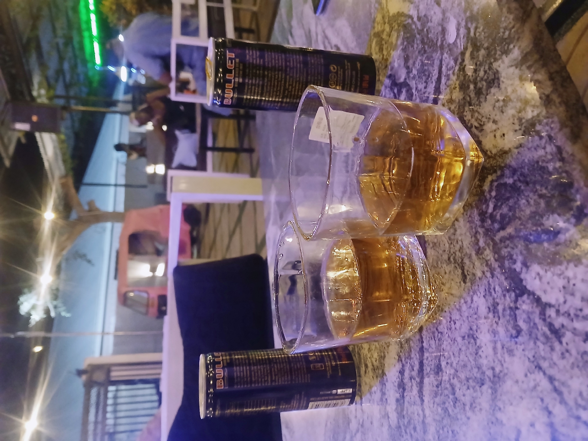 The image shows two glasses filled with a golden liquid, presumably alcoholic beverages, positioned on a marble-patterned surface. A can of "BULLET" energy drink lies near each glass. In the background, there are blurred figures of people seated at tables, suggesting an outdoor dining or bar setting. The presence of lighting and what appears to be a pink vehicle suggests this may be a restaurant or bar area. Jos, Nigeria cannot be determined from the image.