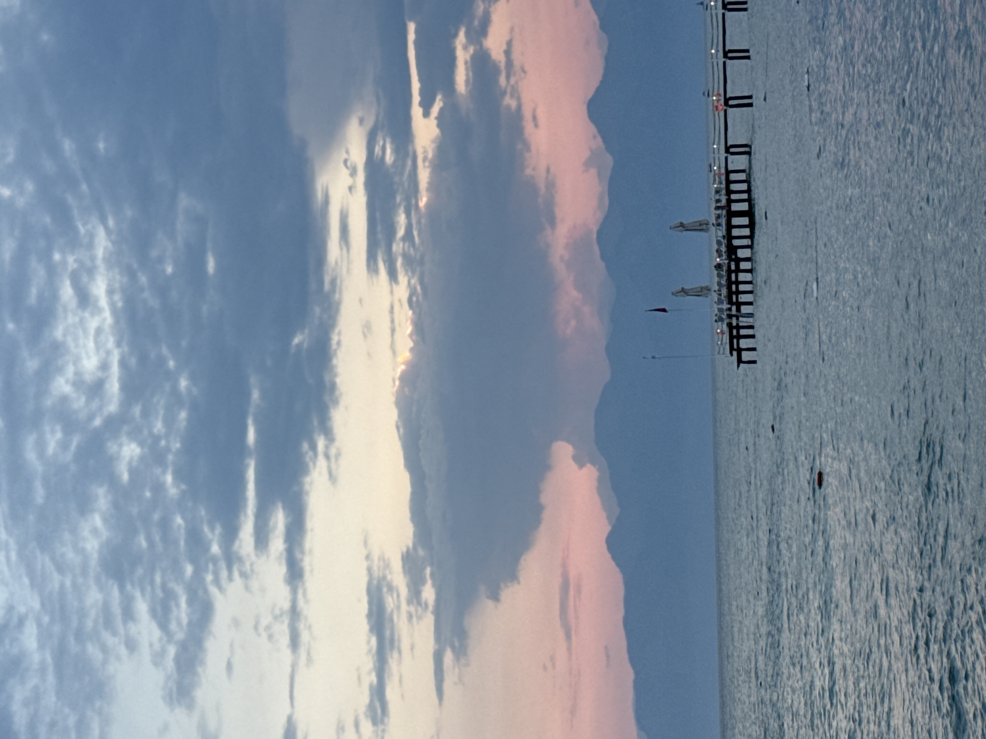 This image depicts a serene seascape at dusk, possibly along the coast of Aksu, Turkey. The foreground is dominated by the calm, blue-grey waters of the sea, with gentle ripples reflecting the fading light. In the middle ground, a wooden pier extends from the right side of the frame, supported by pilings. Along the pier, several beach umbrellas are visible, suggesting a resort or leisure area. A flag flies from a pole on the pier. The background features a range of silhouetted mountains, softened by the twilight haze. The sky above is a canvas of dramatic clouds, with shades of blue, grey, and hints of pink and orange near the horizon, indicating either sunrise or sunset. The overall atmosphere is tranquil and picturesque.
