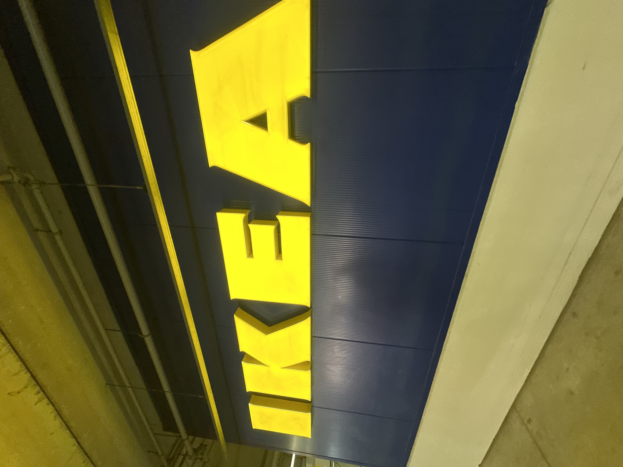 The media file shows a close-up, angled shot of the large, yellow, three-dimensional IKEA logo on a dark blue, corrugated metal facade. The letters spell out "IKEA" vertically. The lighting suggests an indoor, likely commercial, setting, possibly a parking garage or an exterior wall of a building, given the concrete structures and overhead pipes visible in the yellow-tinted areas. The perspective is from below, looking upwards at the sign. The scene is static, with no activity or people visible. The overall impression is a clear, well-lit view of the iconic brand signage. The yellow color of the text stands out vividly against the dark blue background. There are no indications of time of day or weather conditions, as the environment appears to be controlled and indoors or under cover. The location context provided is Alfafar, Spain, which is consistent with the presence of an IKEA store.