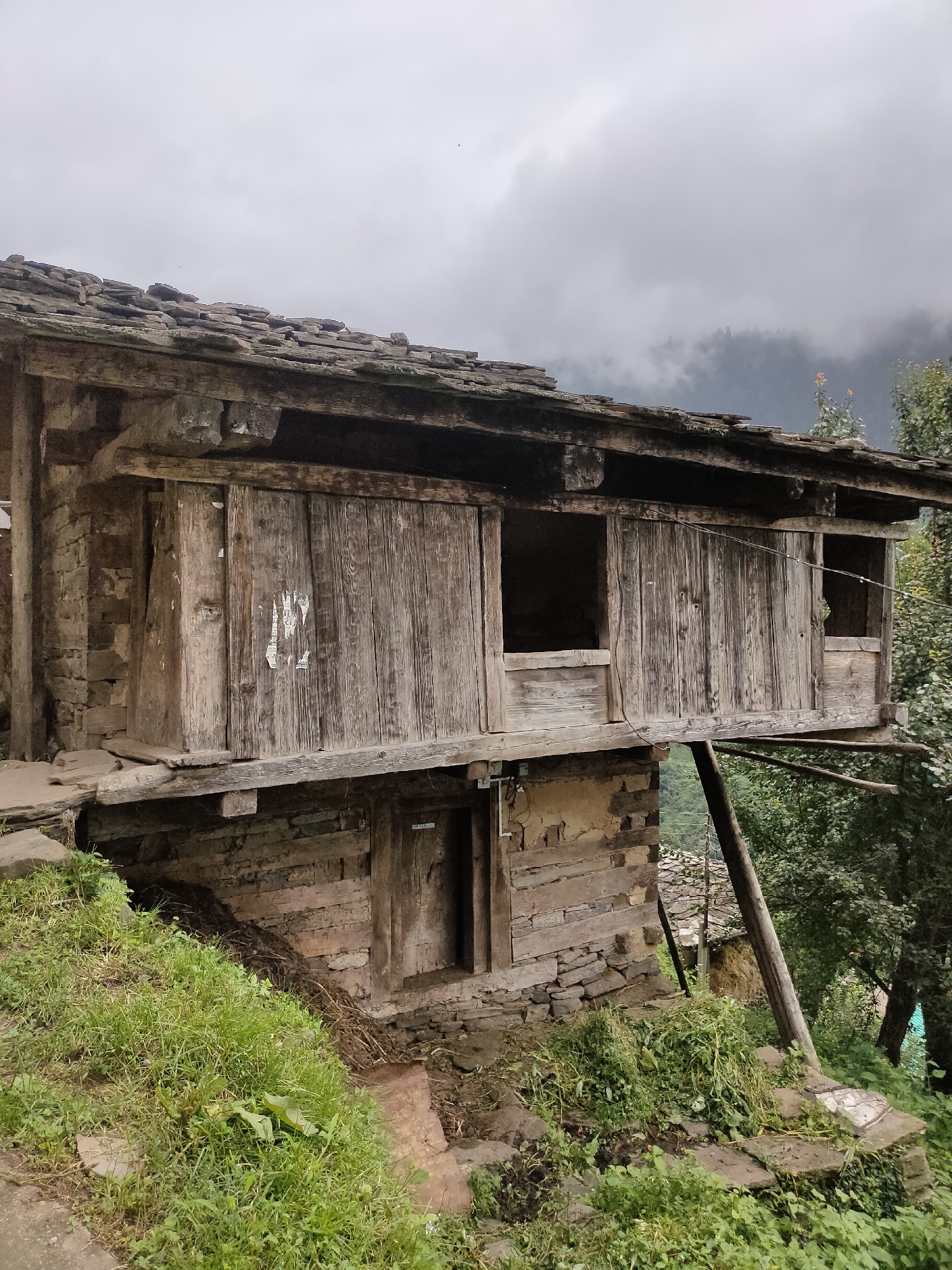 The image depicts a rustic, multi-story wooden and stone structure in Damadi, India. The building appears aged, with weathered wooden planks forming the walls and sliding doors on the upper level. Stone masonry is visible on the lower sections, integrating with the natural slope of the terrain. Green grass and vegetation surround the base of the building, indicating an outdoor, possibly rural or mountainous, setting. The sky above is overcast, suggesting a cloudy or misty day. No people or specific activities are visible in the image. The architecture suggests a traditional style, possibly a dwelling or storage unit.