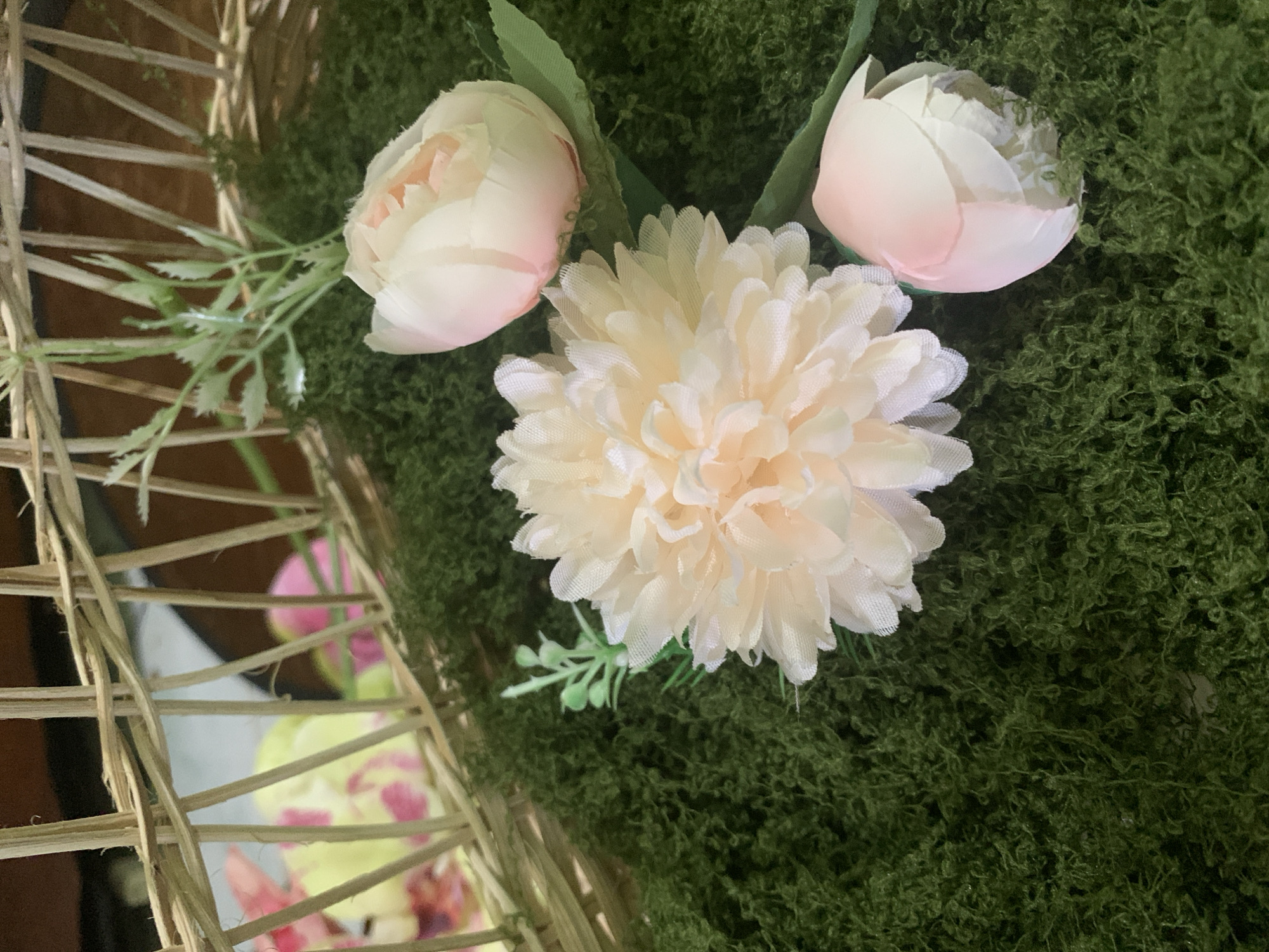 The image depicts a close-up, high-angle view of artificial flowers and greenery arranged within a basket. The primary focus is on three pale pink and cream-colored artificial flowers, one large and fully bloomed, and two smaller buds. The flowers are nestled amongst a bed of textured green material, resembling moss. A portion of a light-colored wicker basket with vertical supports is visible in the upper left quadrant, with a blurred background of what appear to be pink and yellow orchids. The scene is static and appears to be a decorative arrangement, likely indoors. There are no people or discernible actions occurring. The image does not contain any information that would allow for its location to be specified as Kuala Pembuang, Indonesia.