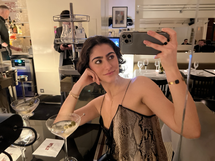A woman takes a selfie in a restaurant. She wears a snakeskin print top, thin strapped black dress, and jewelry. A martini glass sits on the table in front of her, another one is in the background. Other diners are seen in the mirror behind her and to the right. A bartender and other restaurant staff are visible in the background.