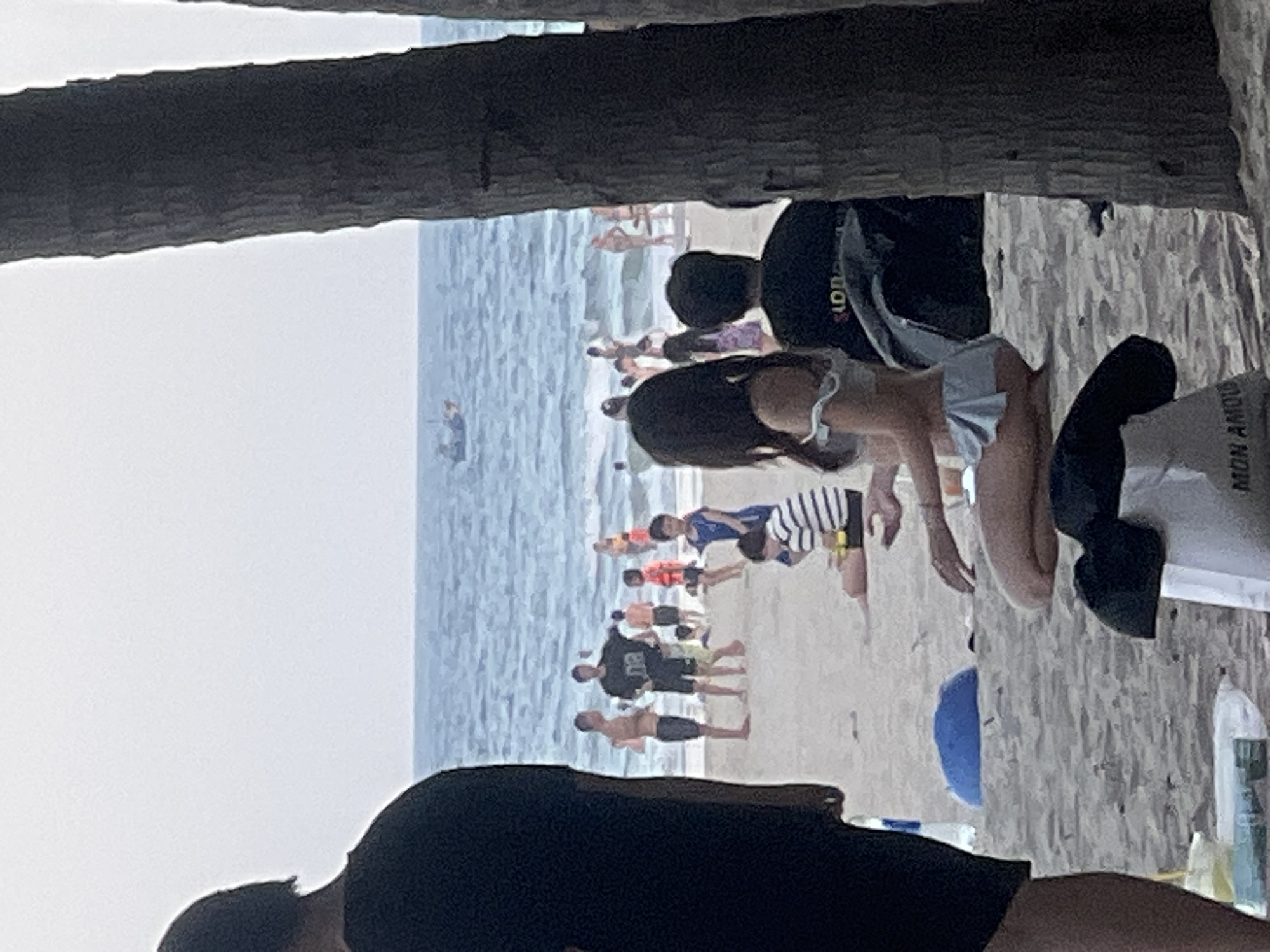 This image depicts a beach scene in Đà Nẵng, Vietnam. The foreground is dominated by the dark silhouettes of what appear to be tree trunks or a wooden structure, creating a frame for the view beyond. The primary subjects are numerous people on a sandy beach and in the shallow ocean waters.
The people are engaged in various beach activities. Some are standing or walking on the sand, while others are in the water. There are several individuals, including children and adults, visible in the distance. Some appear to be swimming or playing in the waves, while others are simply enjoying the beach. The individuals in the foreground are less clearly defined due to the silhouette effect and the image's orientation.
The scene suggests a sunny day, likely in the afternoon, given the brightness of the water and the overall lighting. The ocean appears relatively calm with gentle waves. Among the objects visible are beachwear on the people, and on the right side of the image, there are some plastic bottles and bags.
Notable details include the presence of a small boat in the water in the mid-ground. On the attire of some individuals, there are glimpses of text. For example, on the back of a black shirt worn by a person in the mid-ground, the text "EU" is visible. Another white shirt on the right side of the image has text that partially reads "MON AMOU." The overall atmosphere appears relaxed and recreational, typical of a beach day. The orientation of the image is rotated, with the horizon appearing vertically.