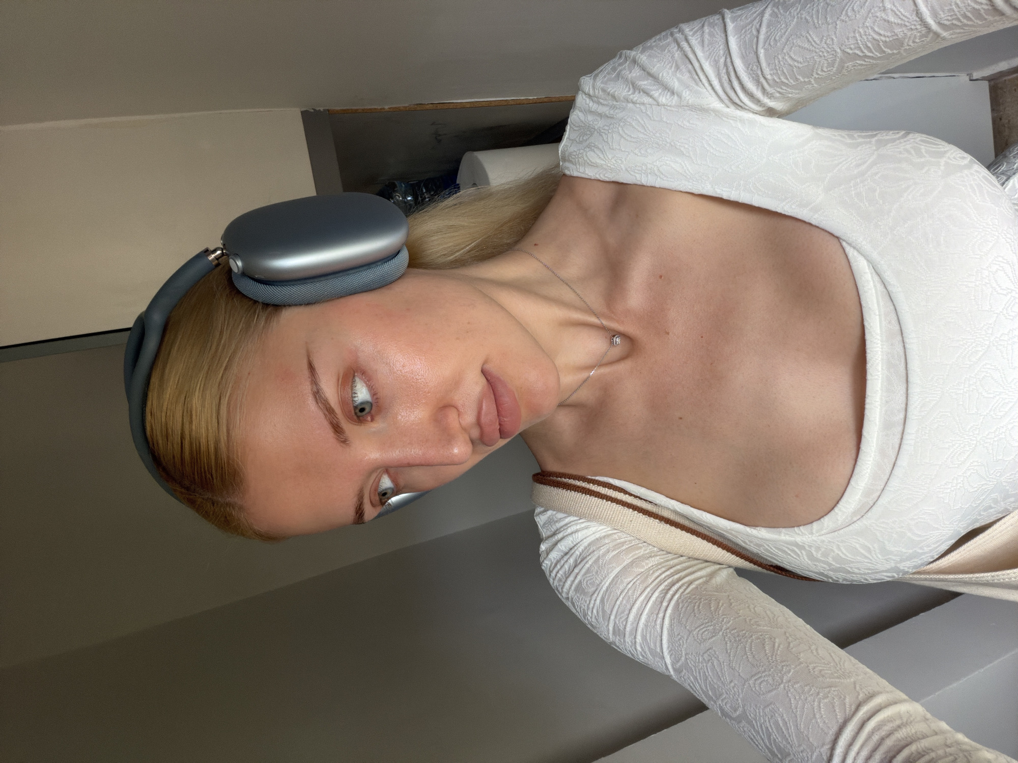 A fair-skinned woman with blonde hair and light blue eyes is wearing gray headphones and a white patterned top. She is also wearing a delicate silver necklace. The background appears to be an interior space with light-colored walls. There is no indication of a specific location in Belgrade, Serbia.